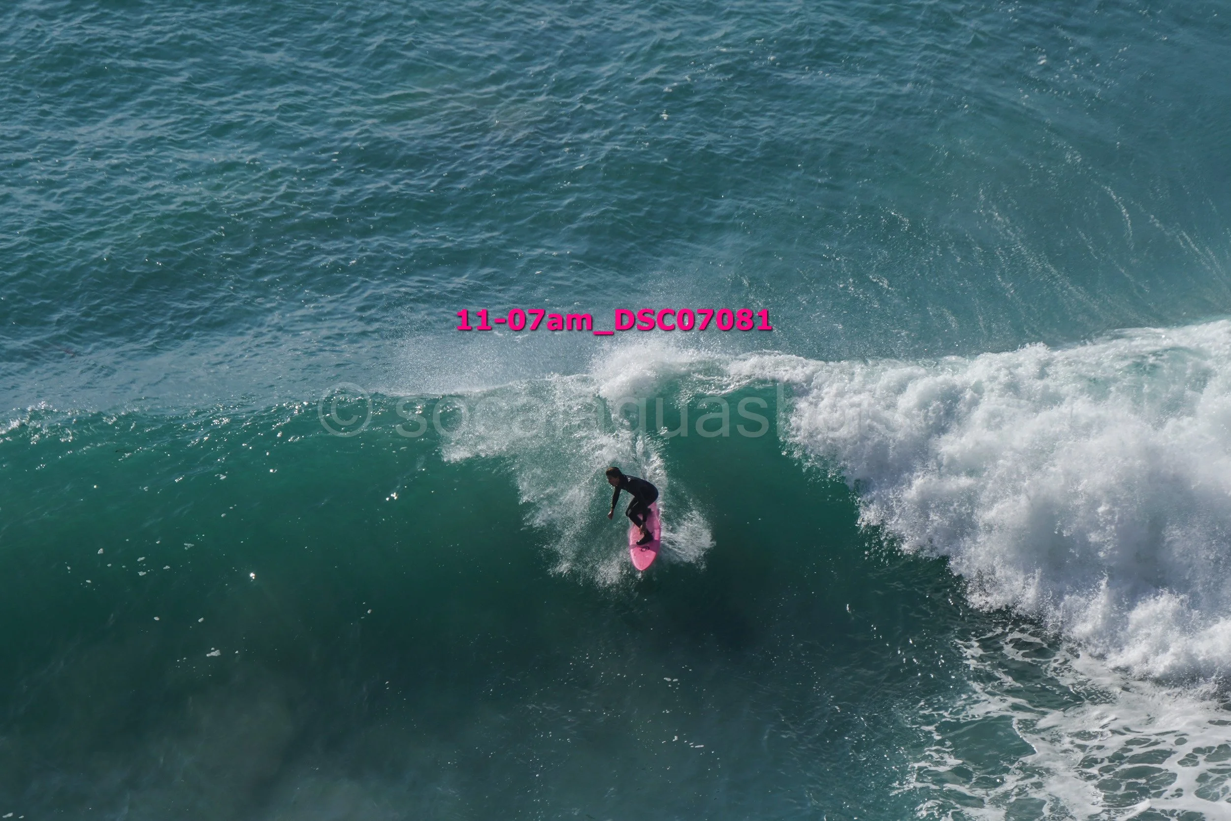 A person surfing on a pink surfboard on a large wave in the ocean with clear blue water.