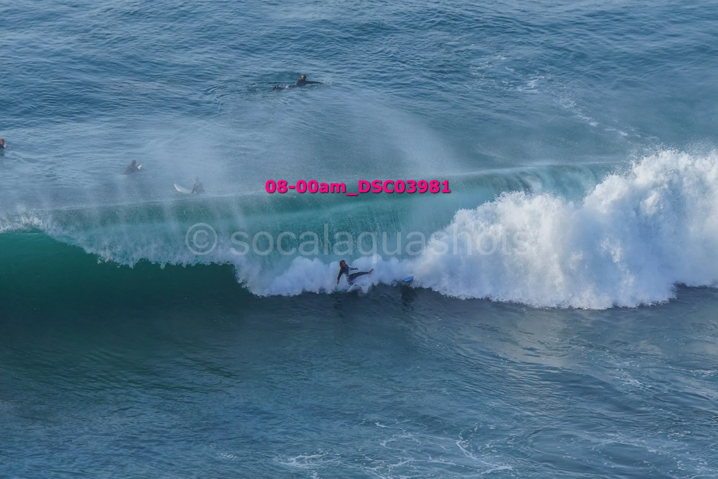 Surfer falling off a large wave in the ocean, with several people in the water in the background.