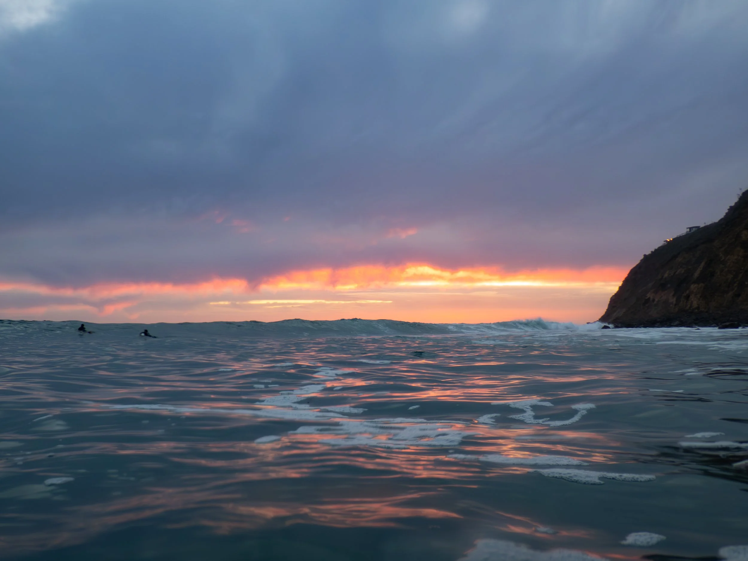 A sunset over the ocean with a cloudy sky, small waves, and a cliff on the right side, with a few surfers in the water.