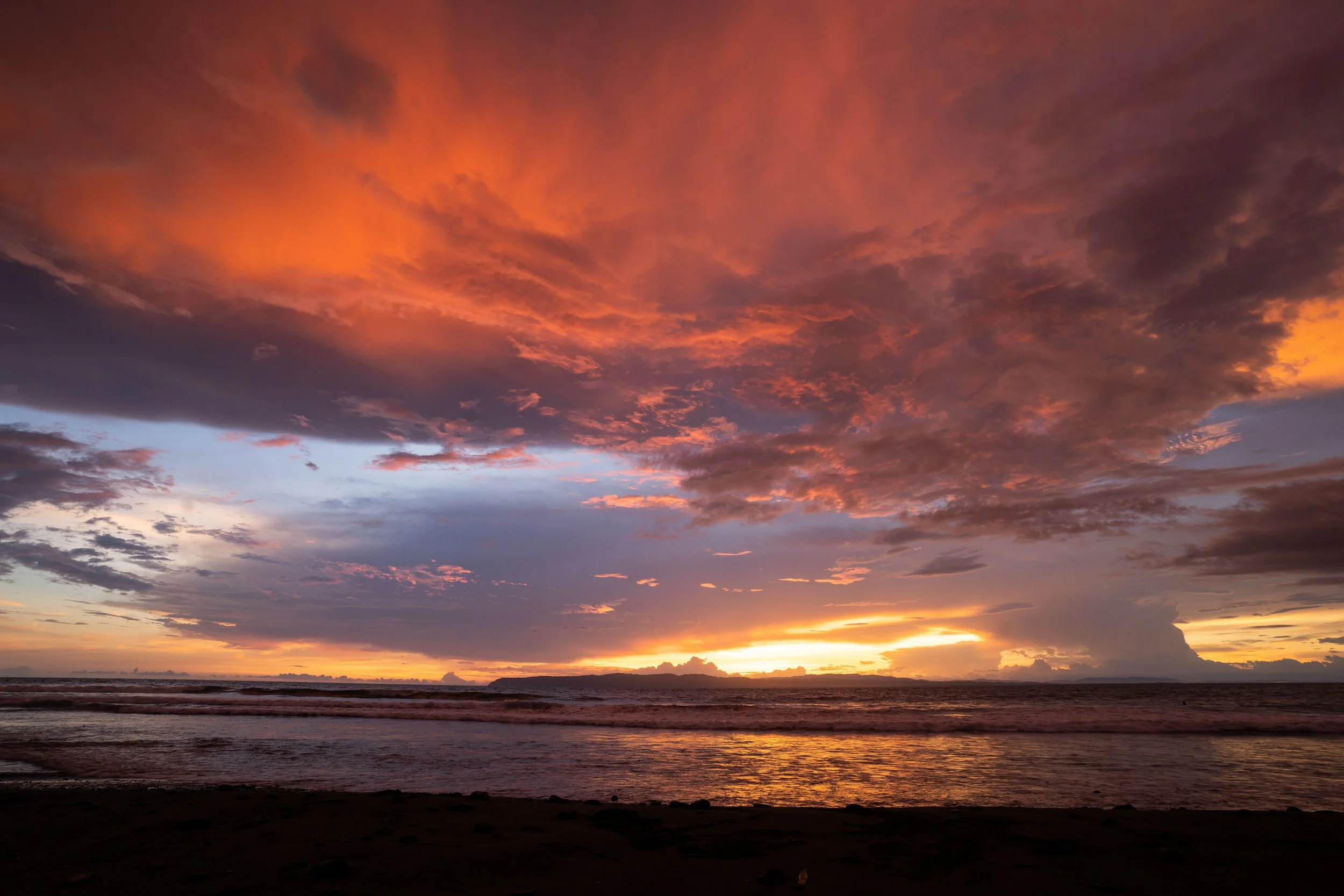 Sunset over the ocean with colorful clouds in the sky, partly cloudy weather, and the shoreline in the foreground.