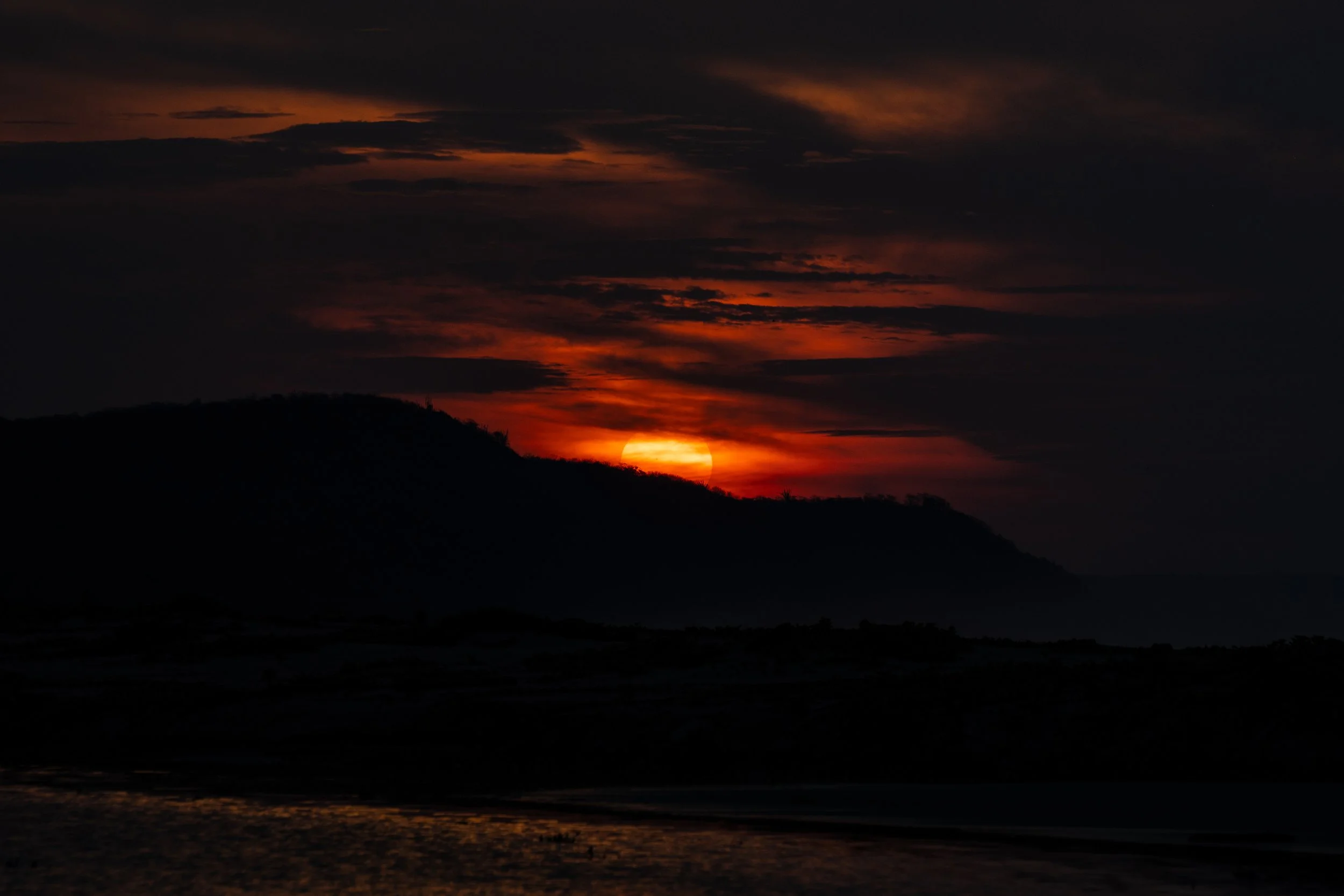 Sunset or sunrise over a hilly landscape with orange and dark clouds in the sky and a reflection on the water.