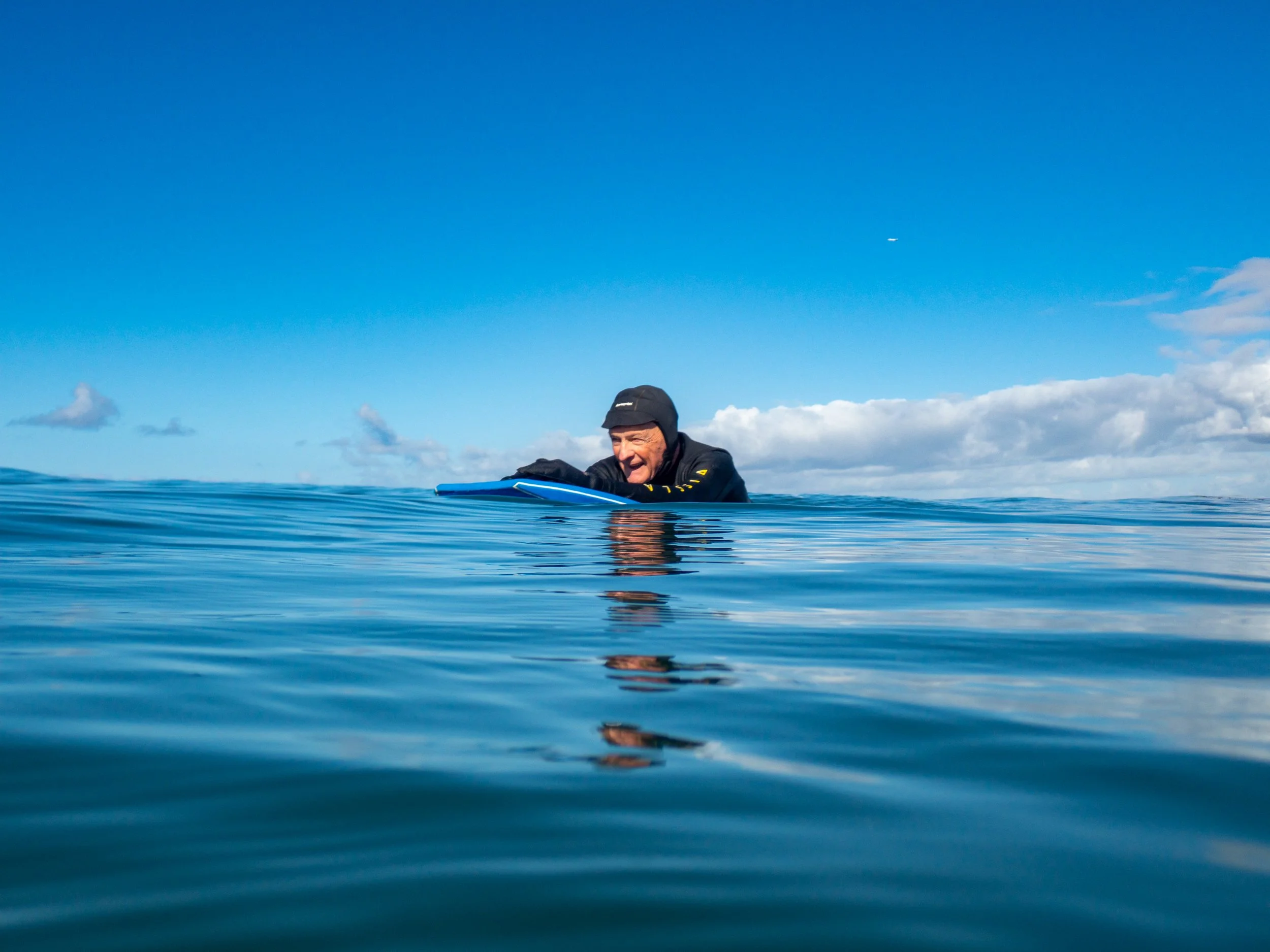 A man in a wetsuit and hood leaning on a surfboard in calm ocean water under a partly cloudy sky.