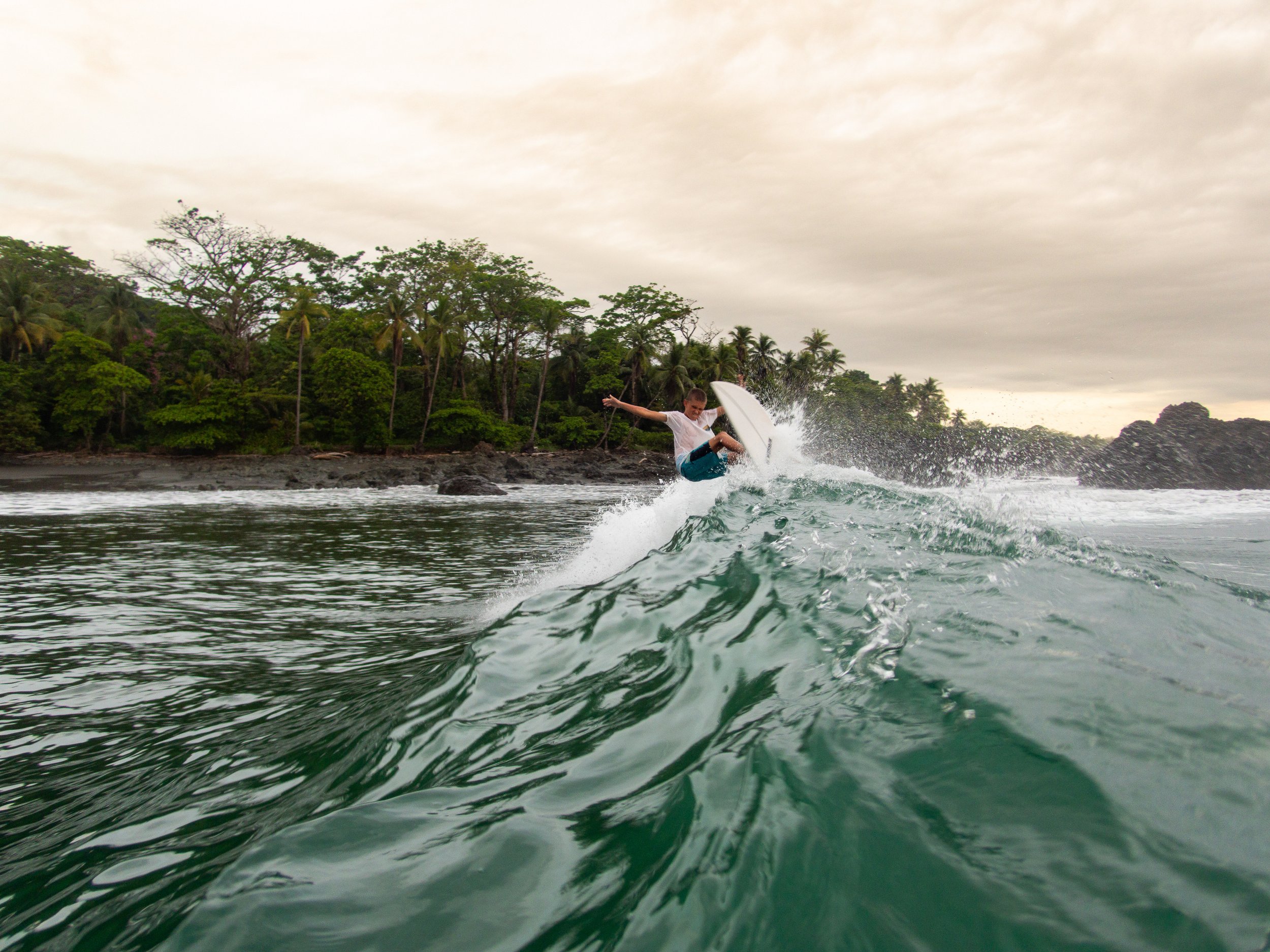Surfer performing maneuver on ocean wave near tropical beach with palm trees under cloudy sky.