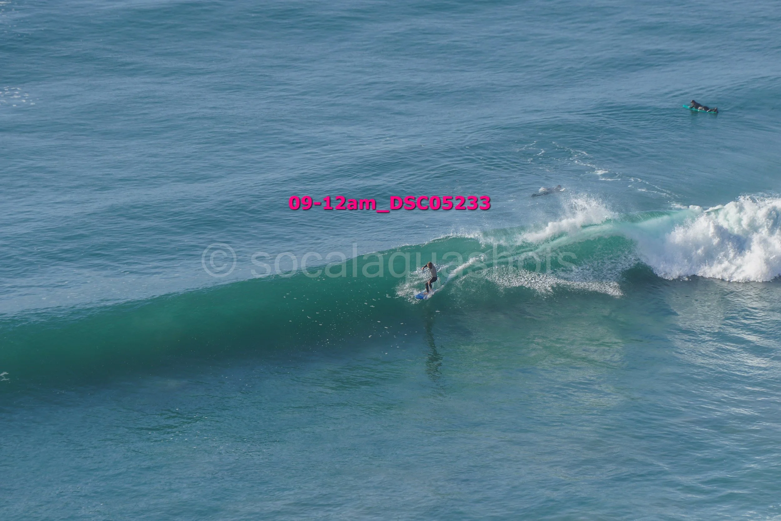 A person surfing on a wave in the ocean, with another surfer in the distance. The water is clear blue, and the surfer is riding the wave near the shoreline.