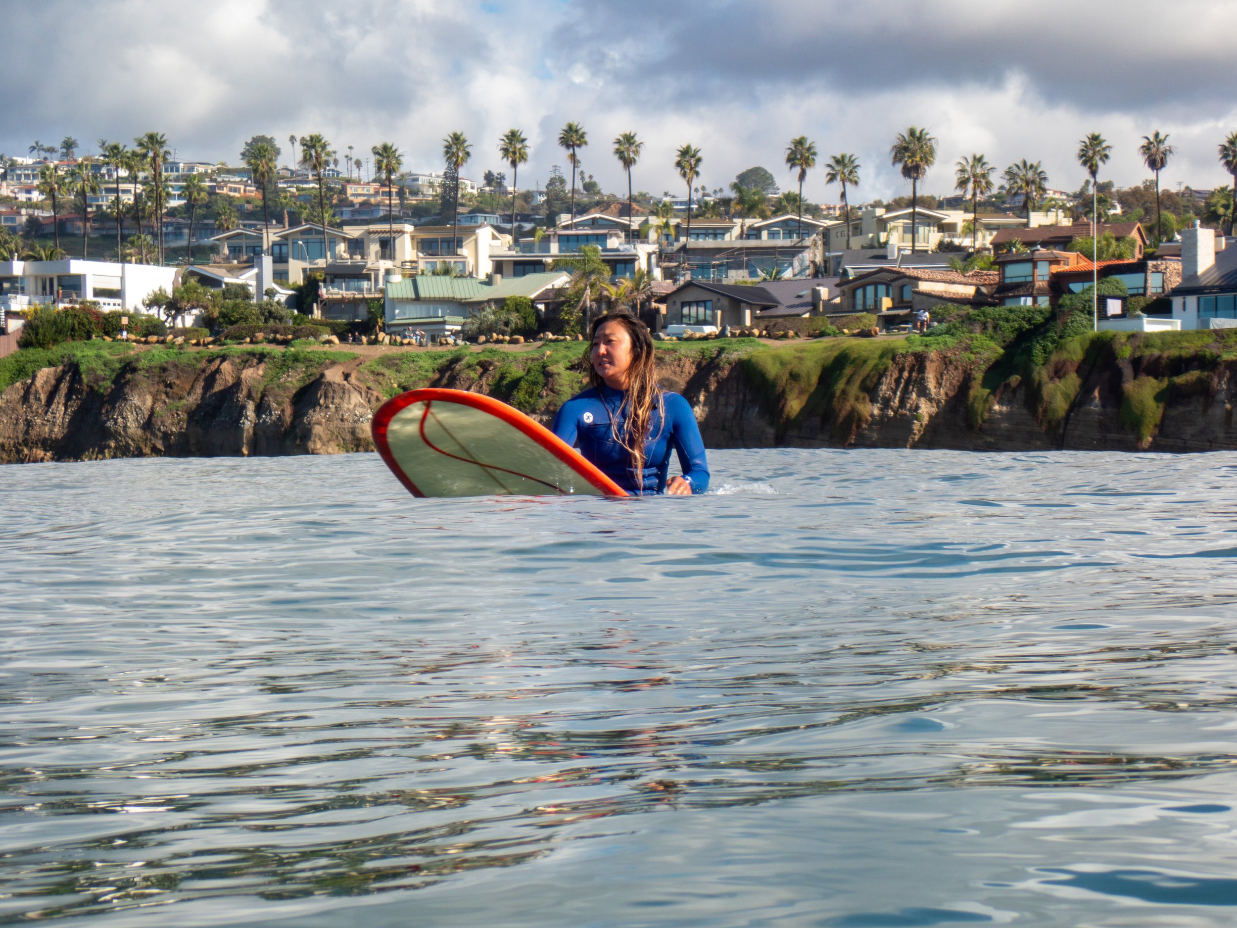 A woman in a blue wetsuit holding a surfboard in the water near a coastal neighborhood with houses and palm trees.