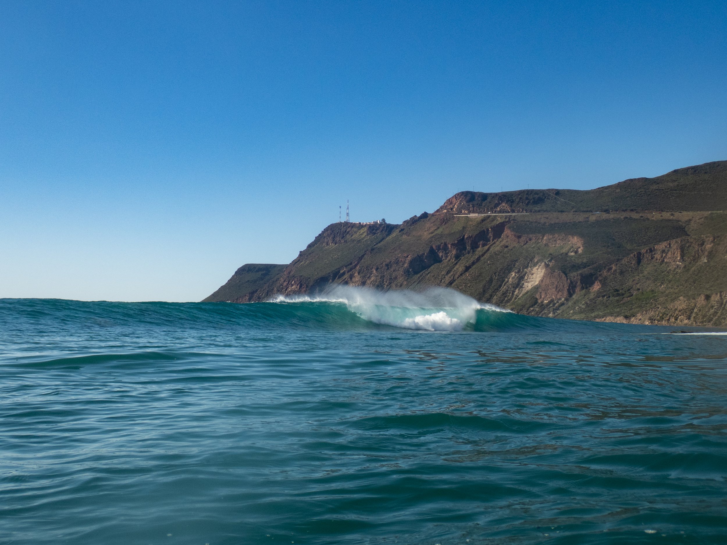 Ocean waves crashing against rocky coastal cliffs under a clear blue sky.