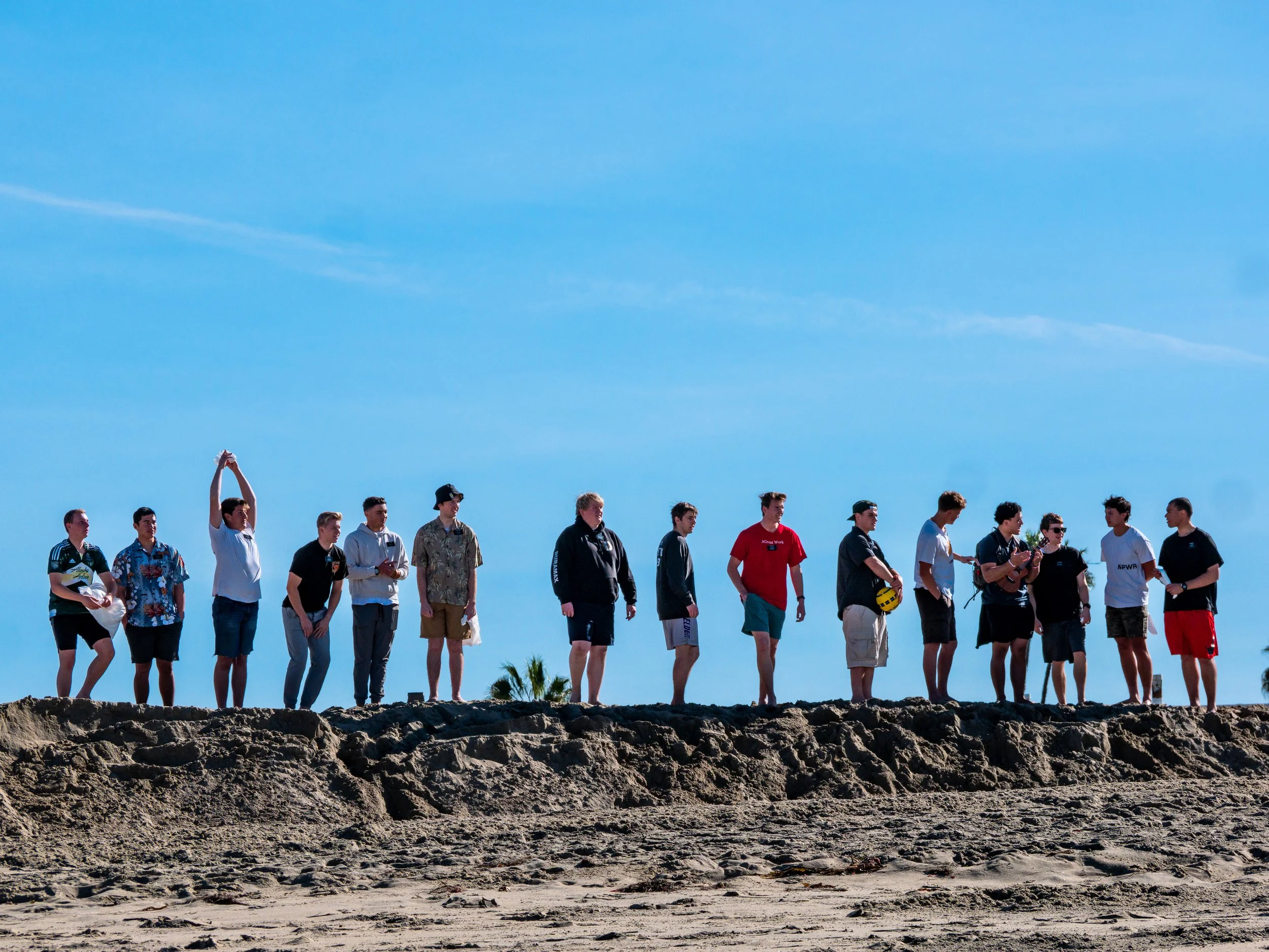 Group of fifteen young people standing on a rocky beach under a clear blue sky, some holding a soccer ball, dressed casually.