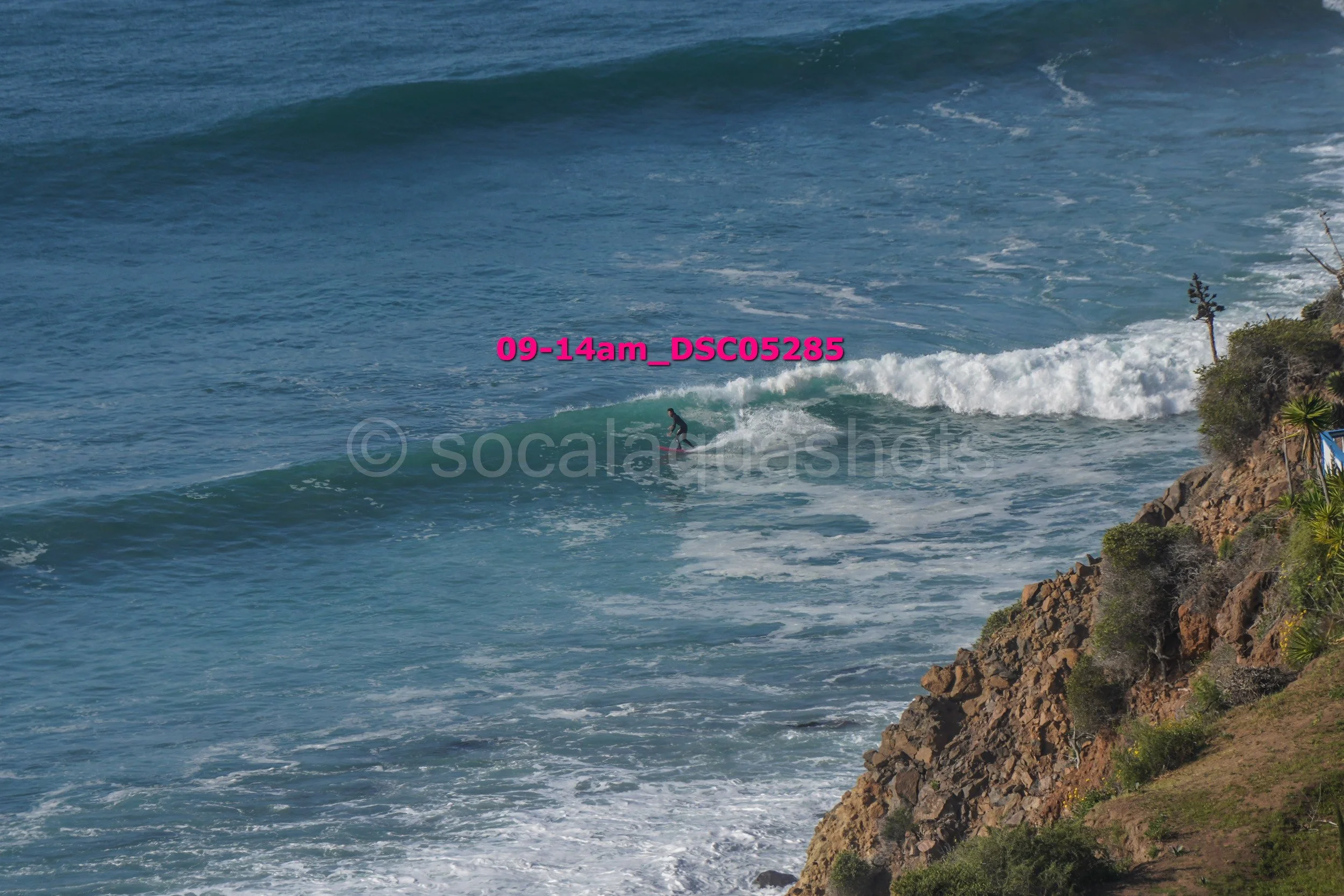 A person surfing on a wave near a rocky coastline with greenery.