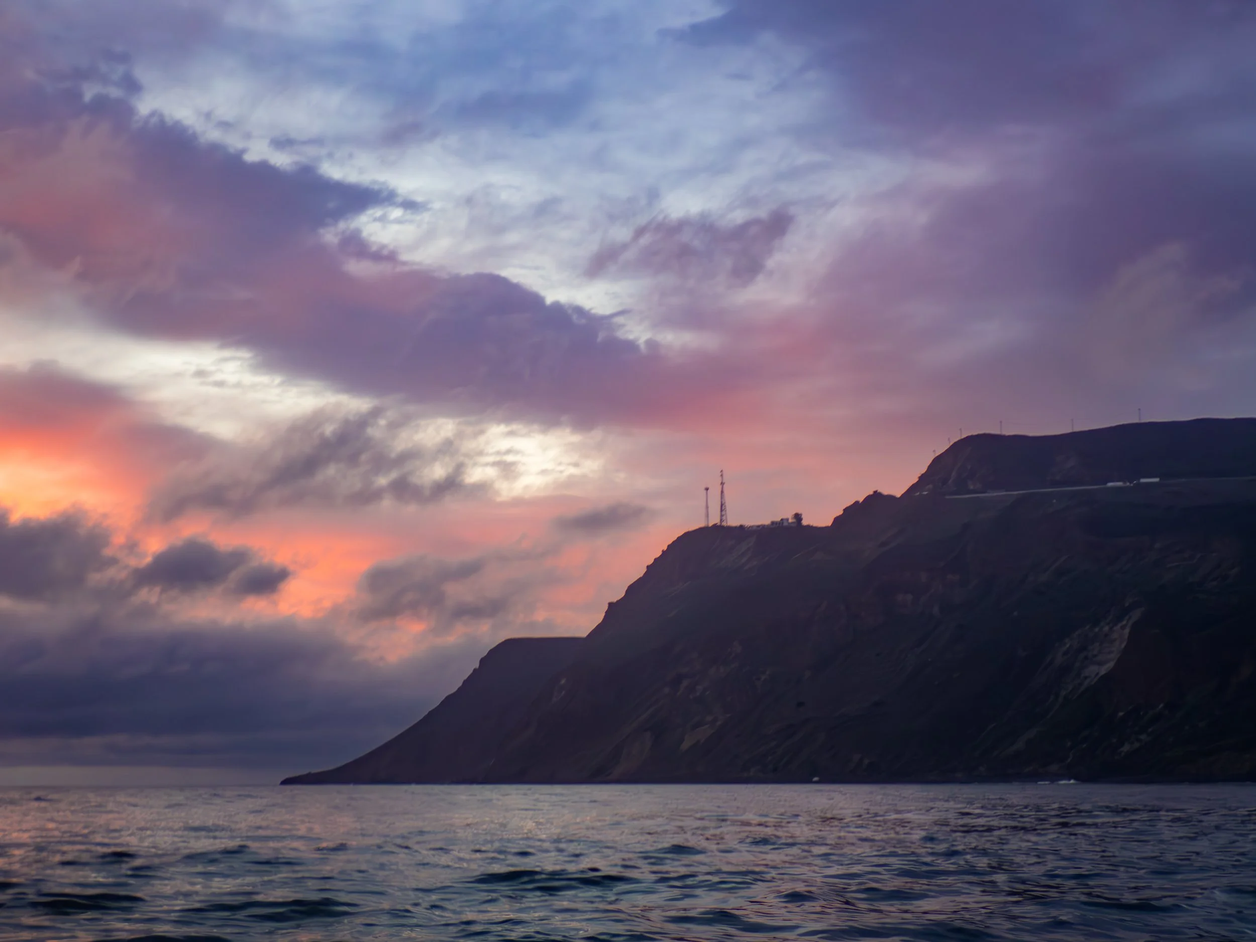 Sunset over the ocean with a cloudy sky and a hillside with communication towers.
