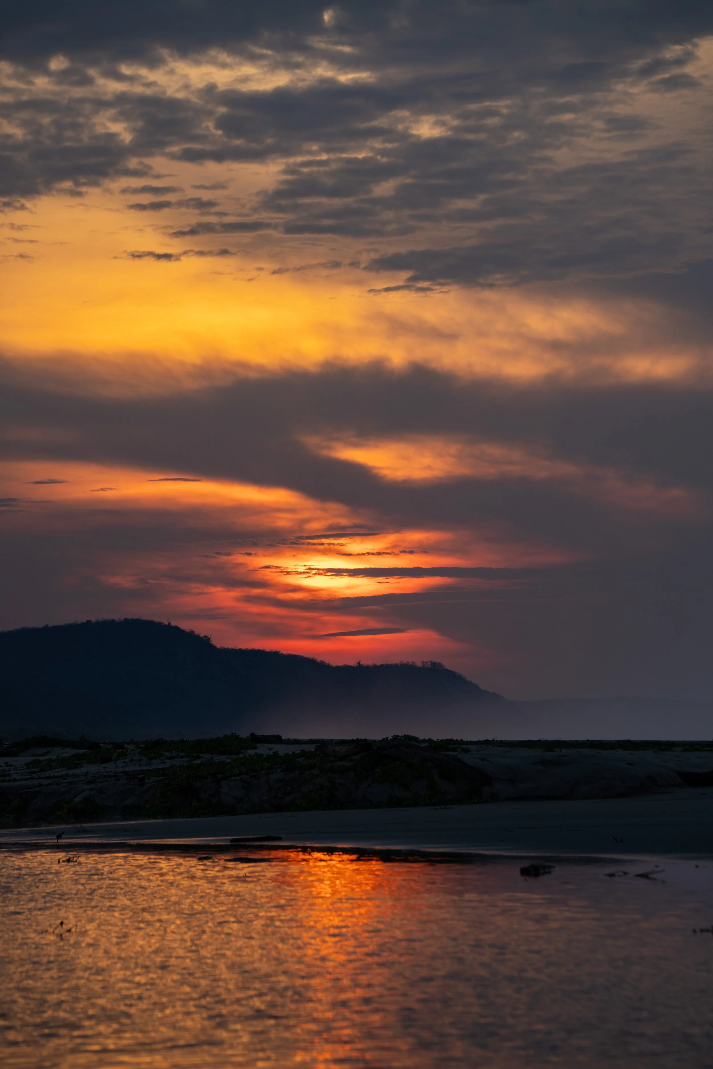 Sunset over mountains with reflection on body of water, dark clouds in sky.