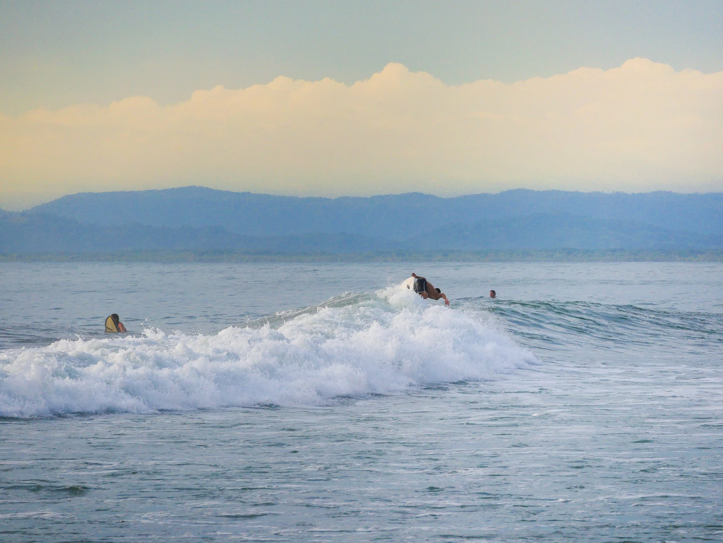 Surfers on waves with mountainous shoreline in background under cloudy sky