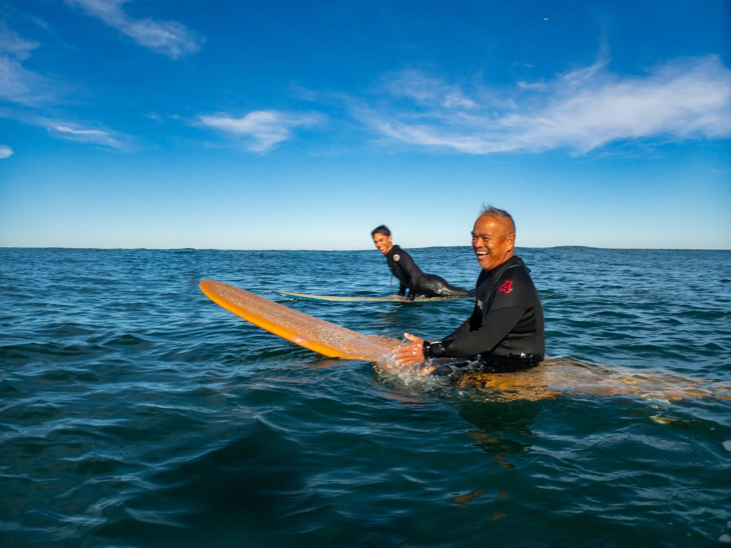 Two men in wetsuits sitting on surfboards in the ocean, smiling, with a clear blue sky above.