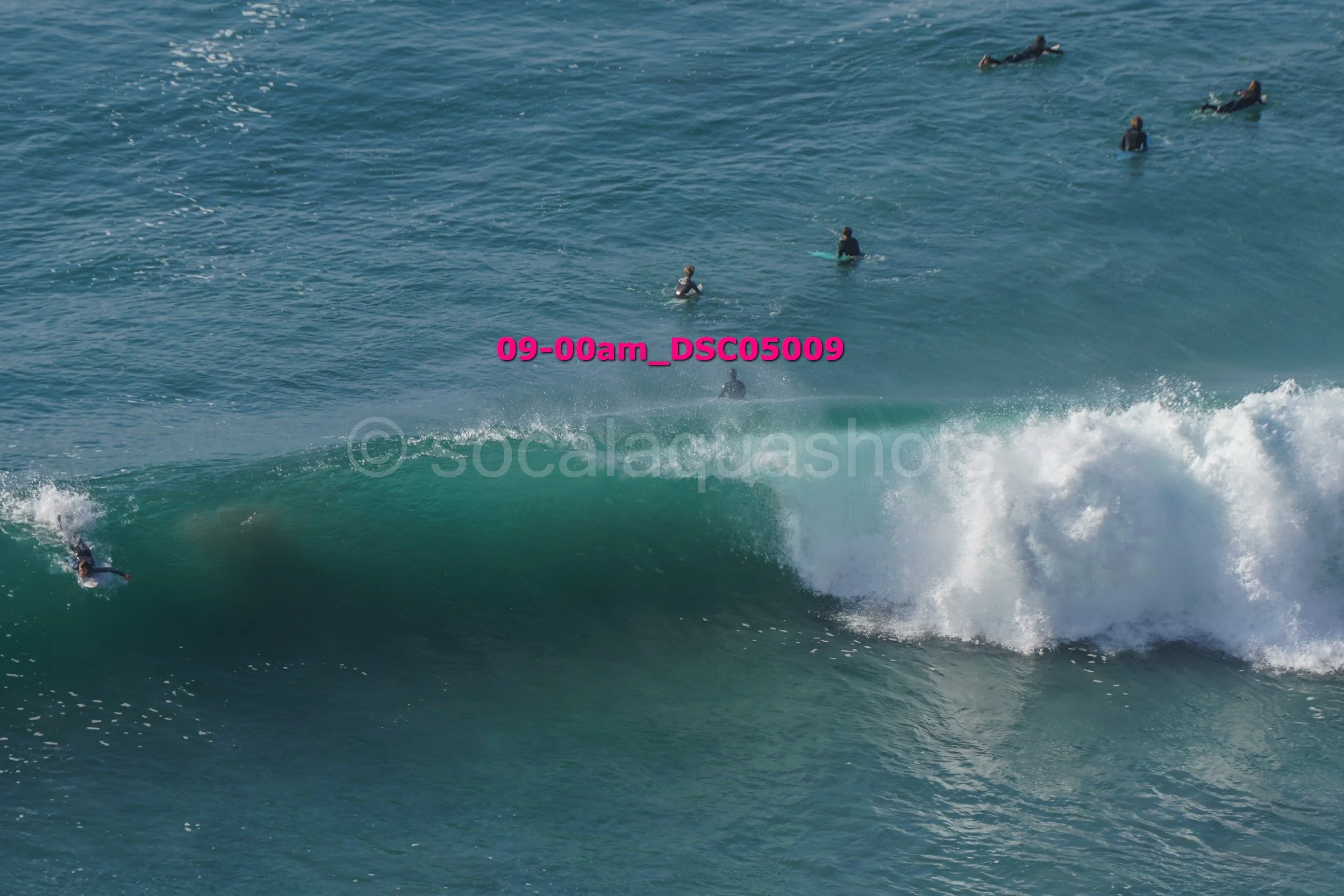 Surfers in the ocean, some riding a wave and others waiting in the water, with a rainbow-colored surfboard visible.