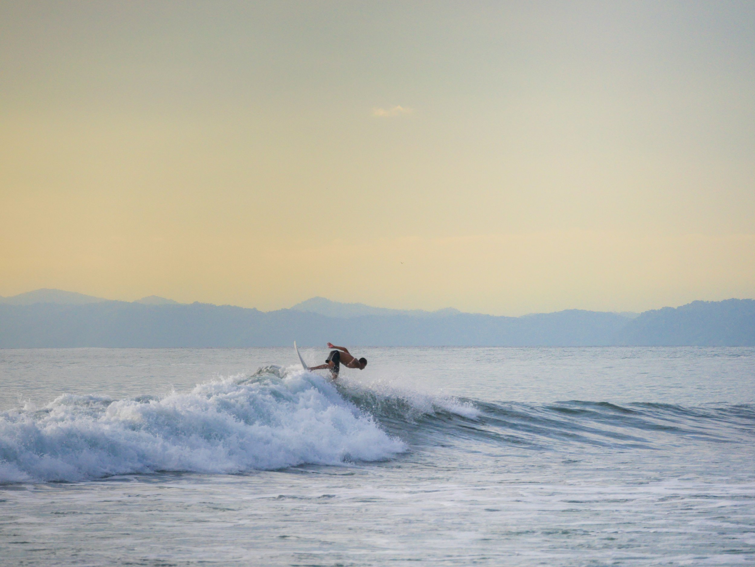 Surfer riding a wave in the ocean with distant hills under a cloudy sky
