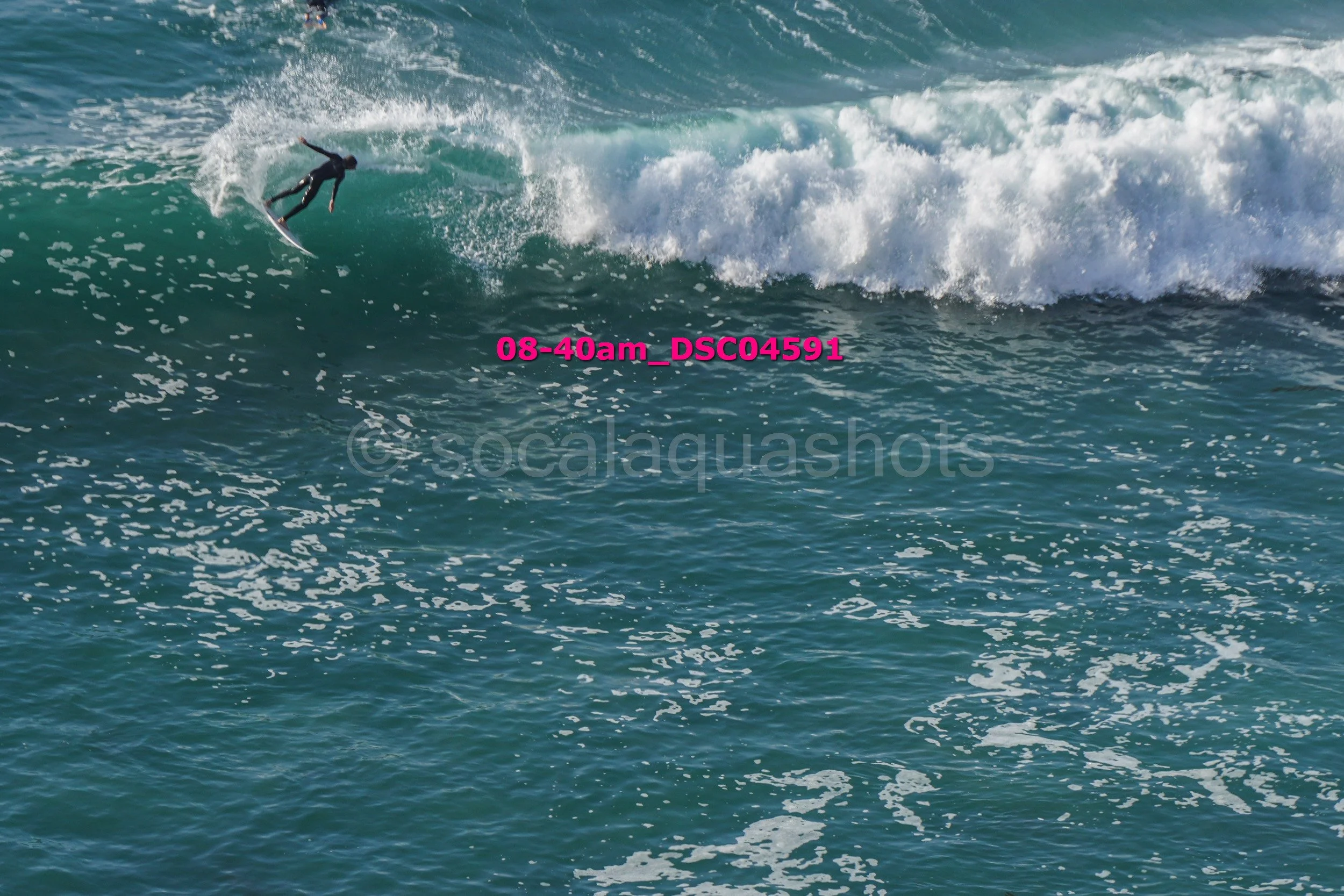 A surfer in a black wetsuit riding a large wave with white foam at the top, seen from above in the ocean.