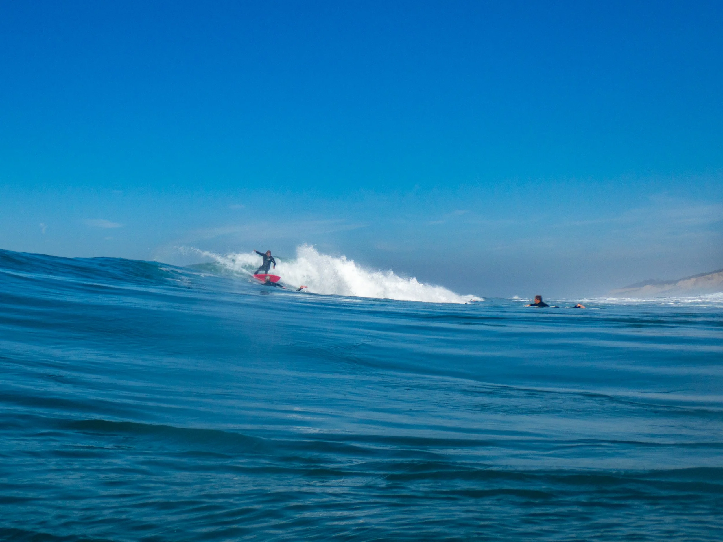 A person surfing on a wave in the ocean with two other people swimming nearby and distant land in the background under a clear blue sky.