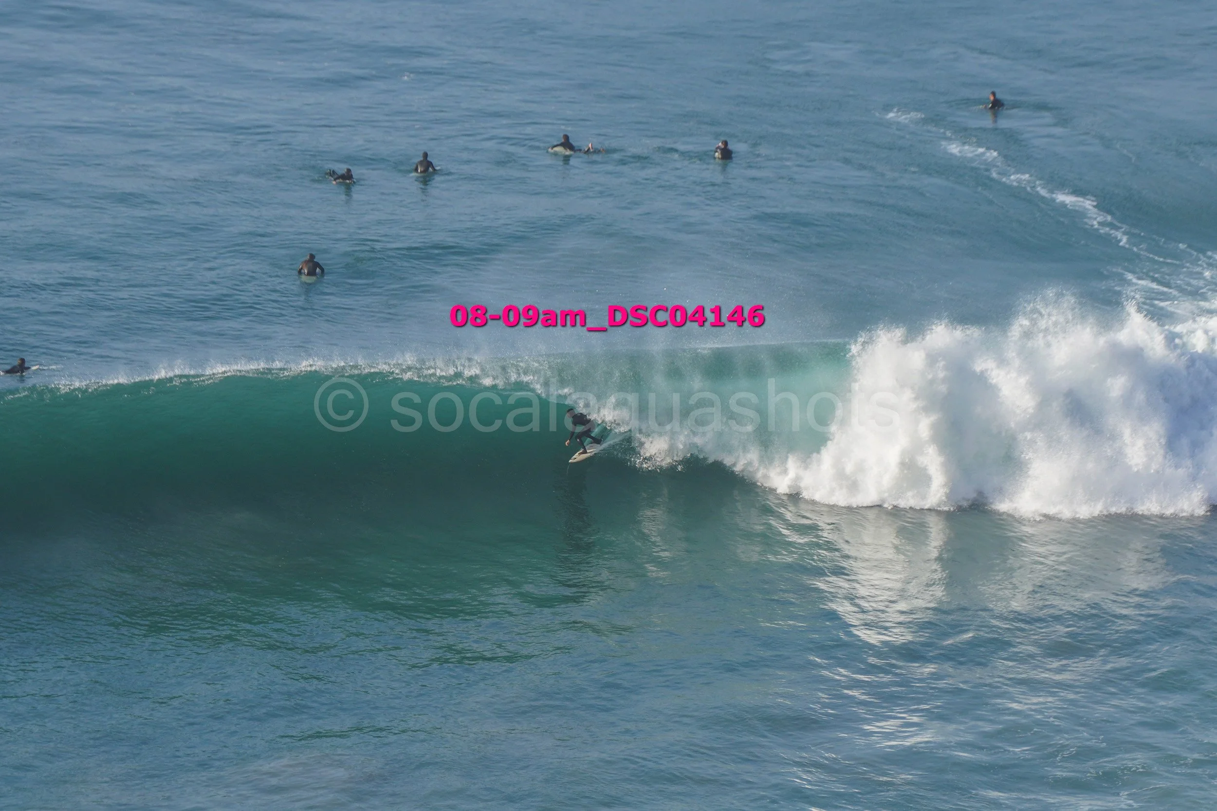 A person surfing on a wave with multiple people swimming and surfing in the background in the ocean.