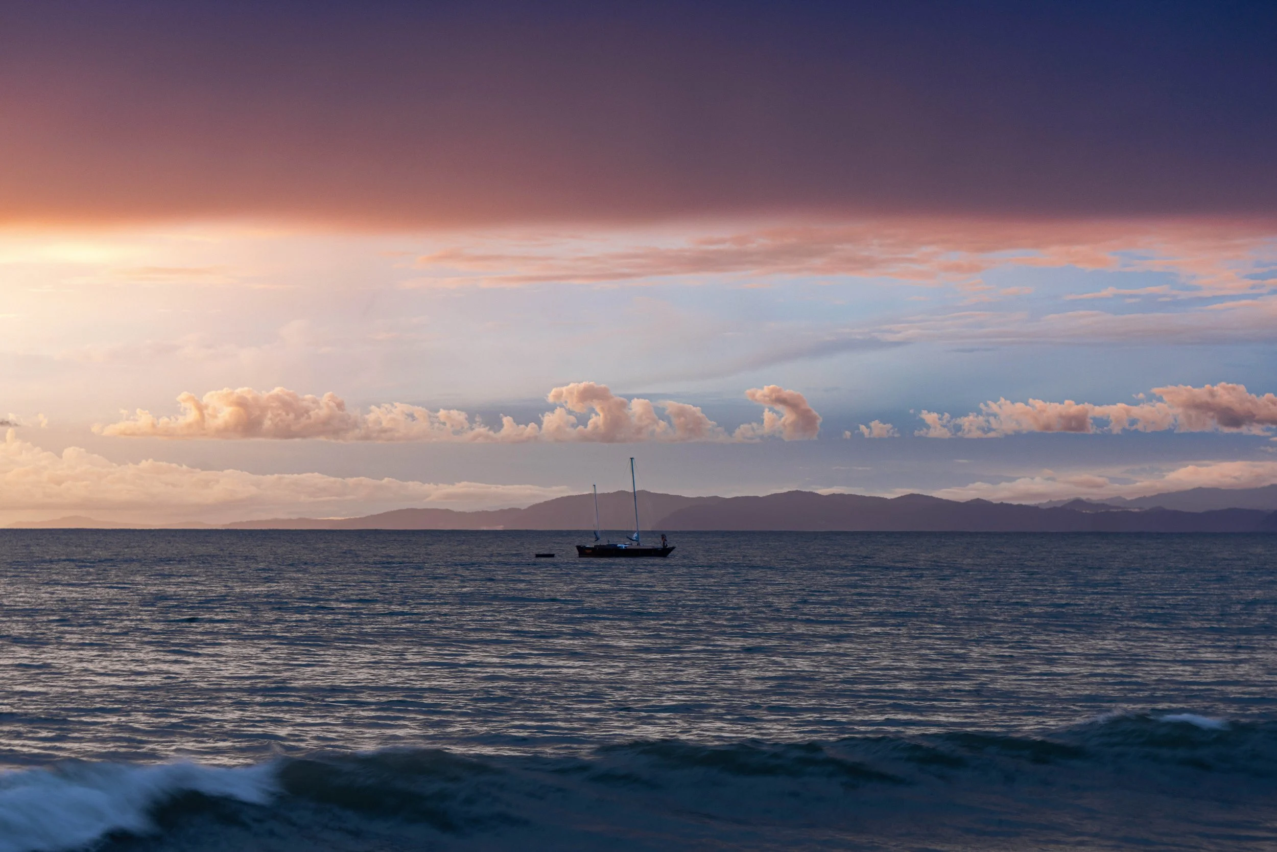A sailboat floating on calm ocean waters at sunset, with a distant mountain range under a sky filled with colorful clouds.