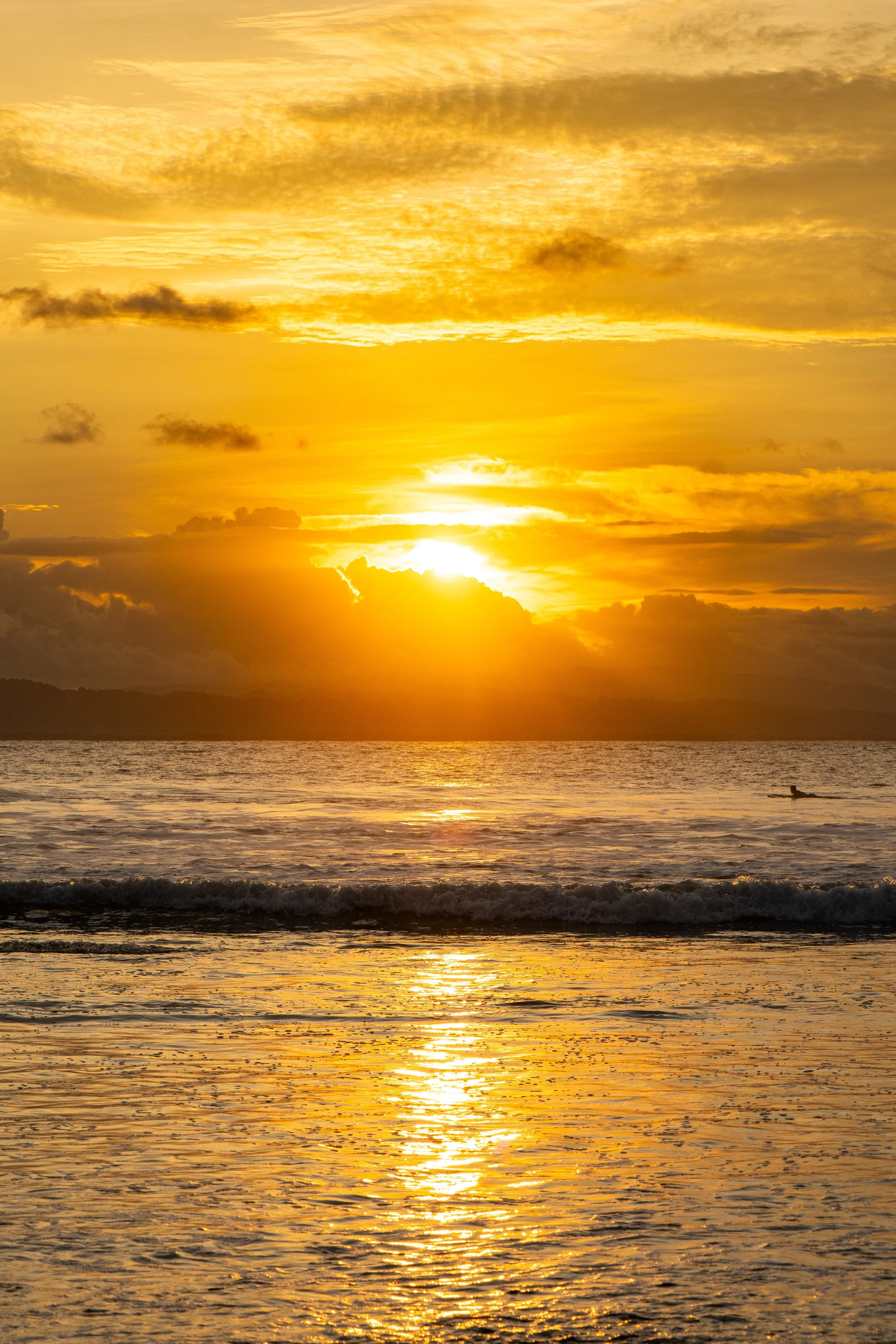 A sunset over the ocean with a bright sun near the horizon, yellow and orange sky, some clouds, and a small boat in the water.