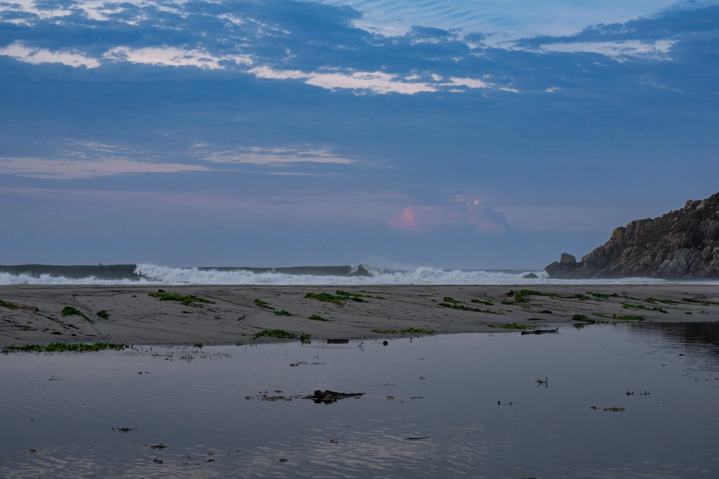 A beach with waves crashing against rocks, a cloudy sky, and tide pools reflecting the sky.
