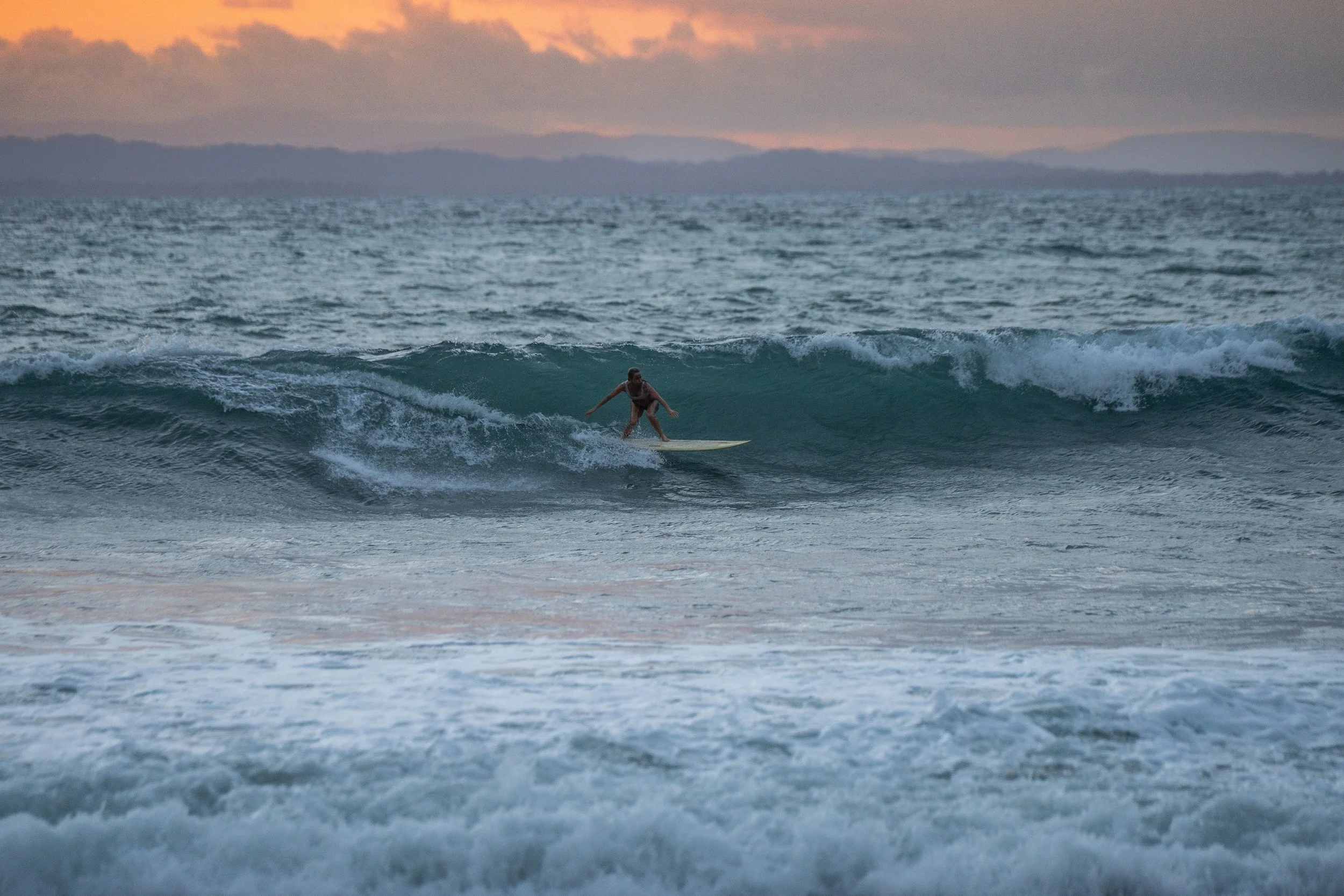A person surfing on a wave in the ocean during sunset with mountains in the background.