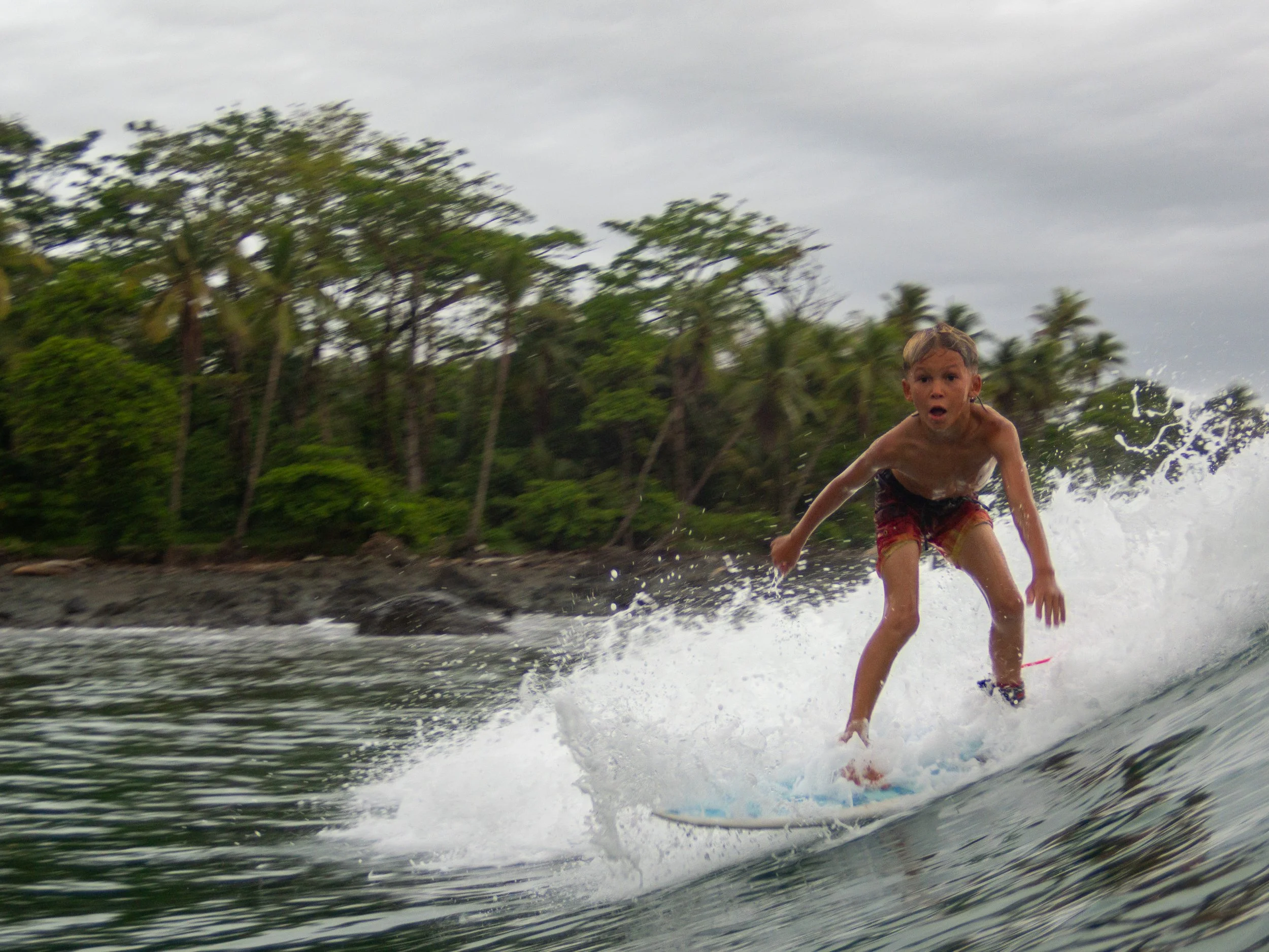 Young boy surfing on a wave near a lush, tropical coastline with trees in the background.