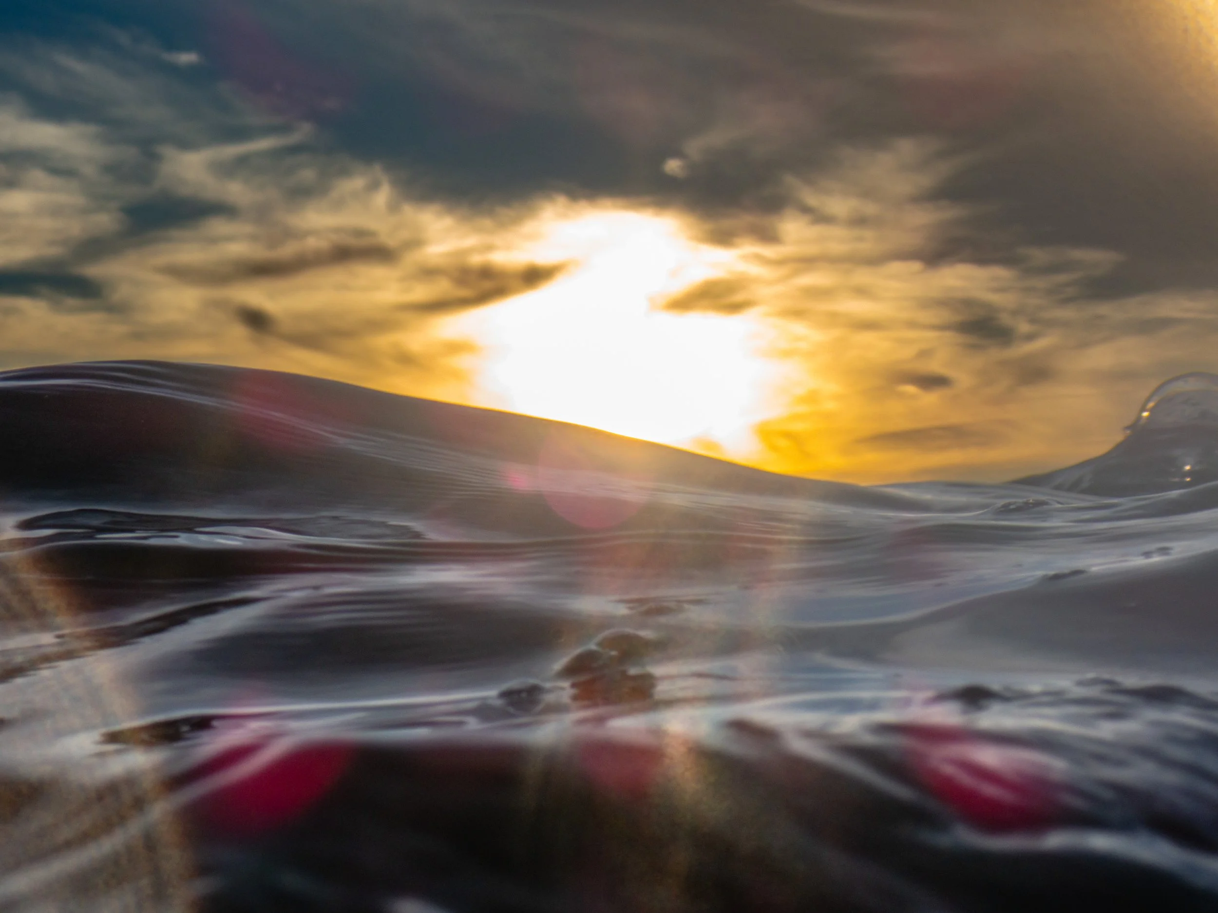 Close-up of ocean waves at sunset with the sun low on the horizon and partly cloudy sky.