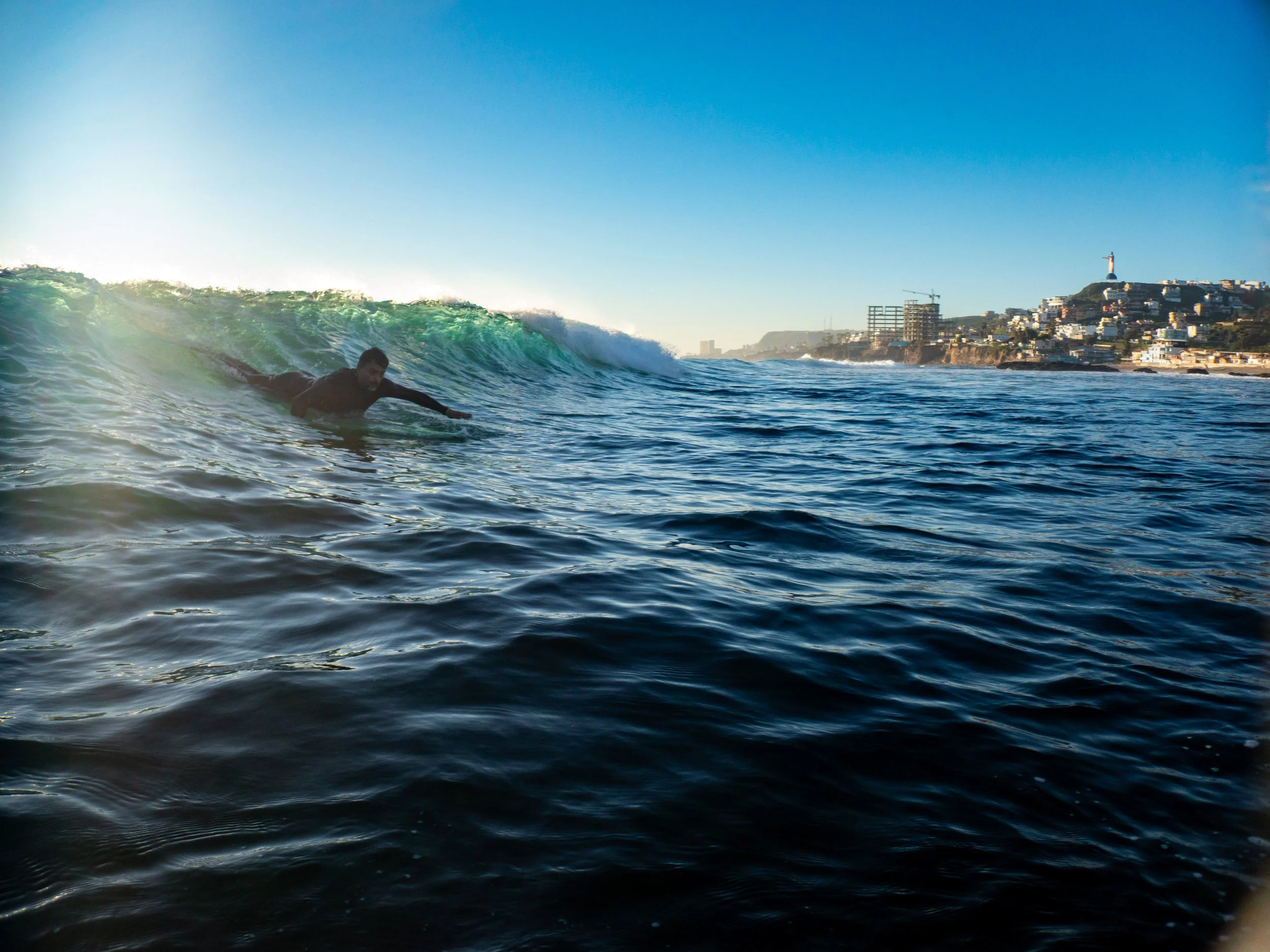 A person surfing on a wave near a city coastline at sunset.
