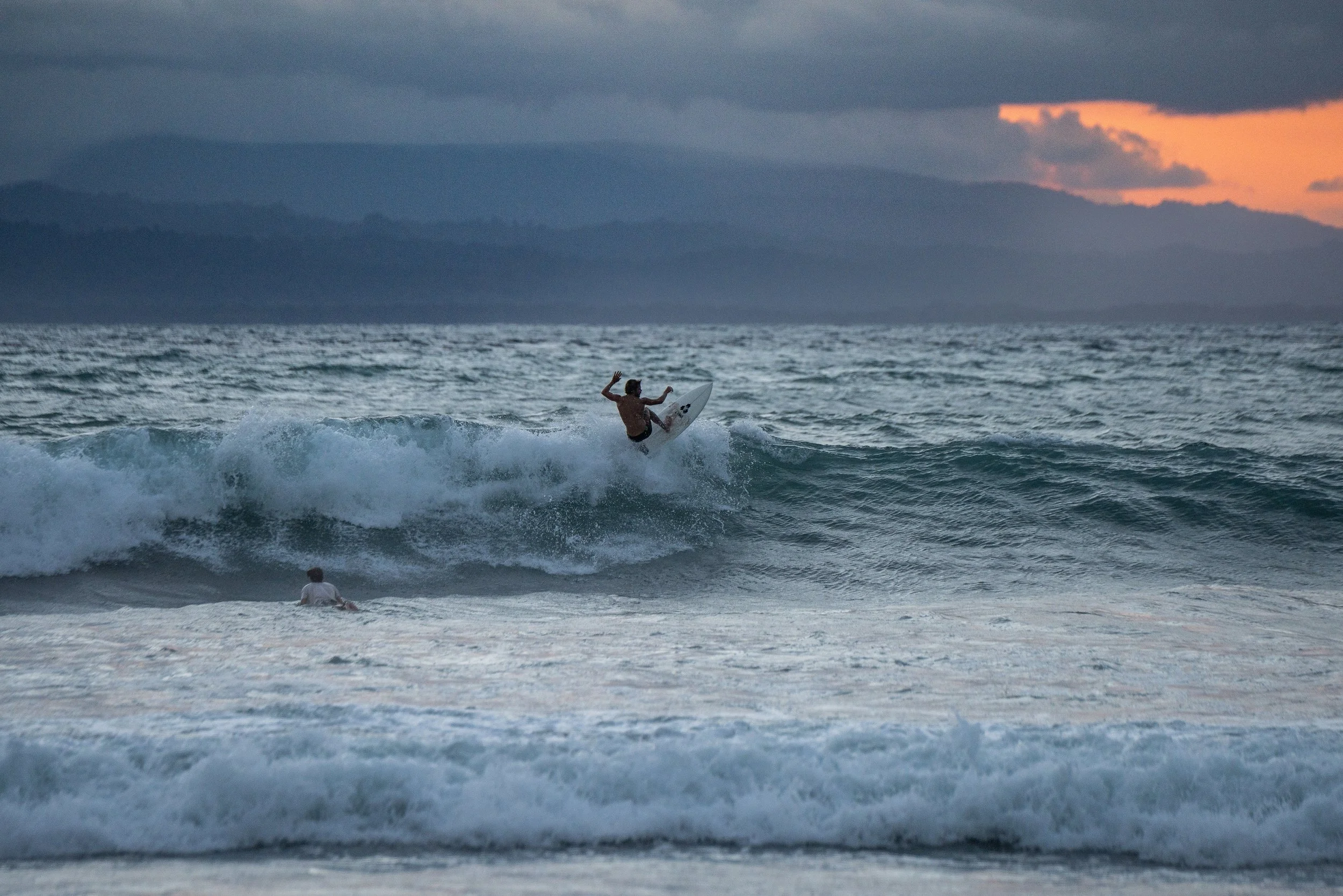 A person surfing on a wave in the ocean during sunset, with dark clouds in the sky and a distant mountain range on the horizon.