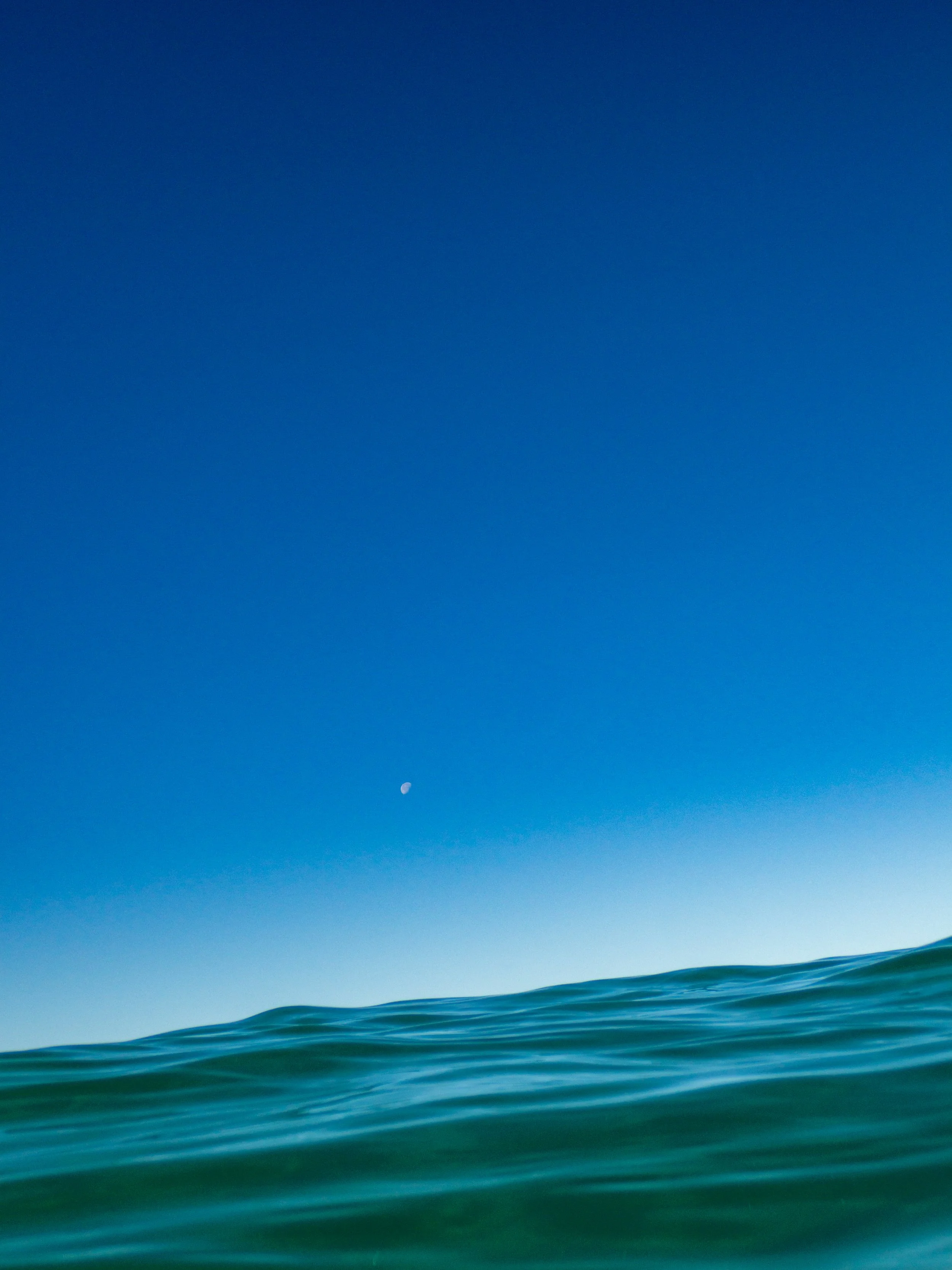 Calm ocean waves with a clear blue sky and a small visible moon.