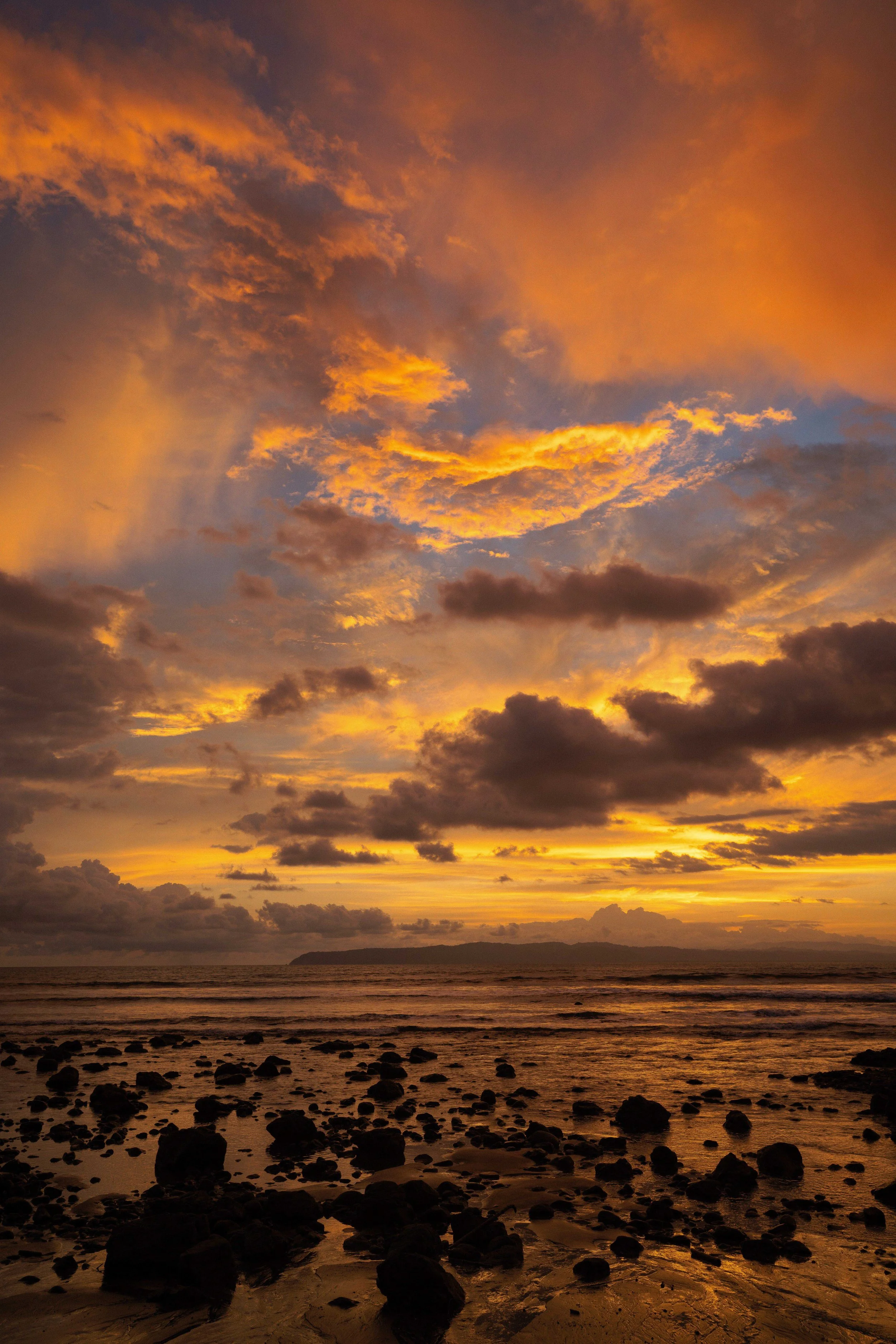 A sunset over the ocean with orange and yellow clouds, a silhouette of a distant island, and rocks on the shore in the foreground.