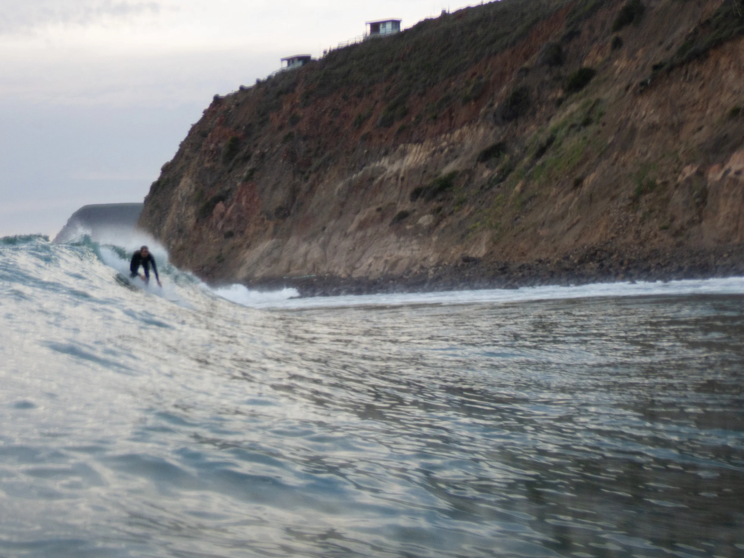 A person surfing in the ocean near a rocky cliff with some houses on top.