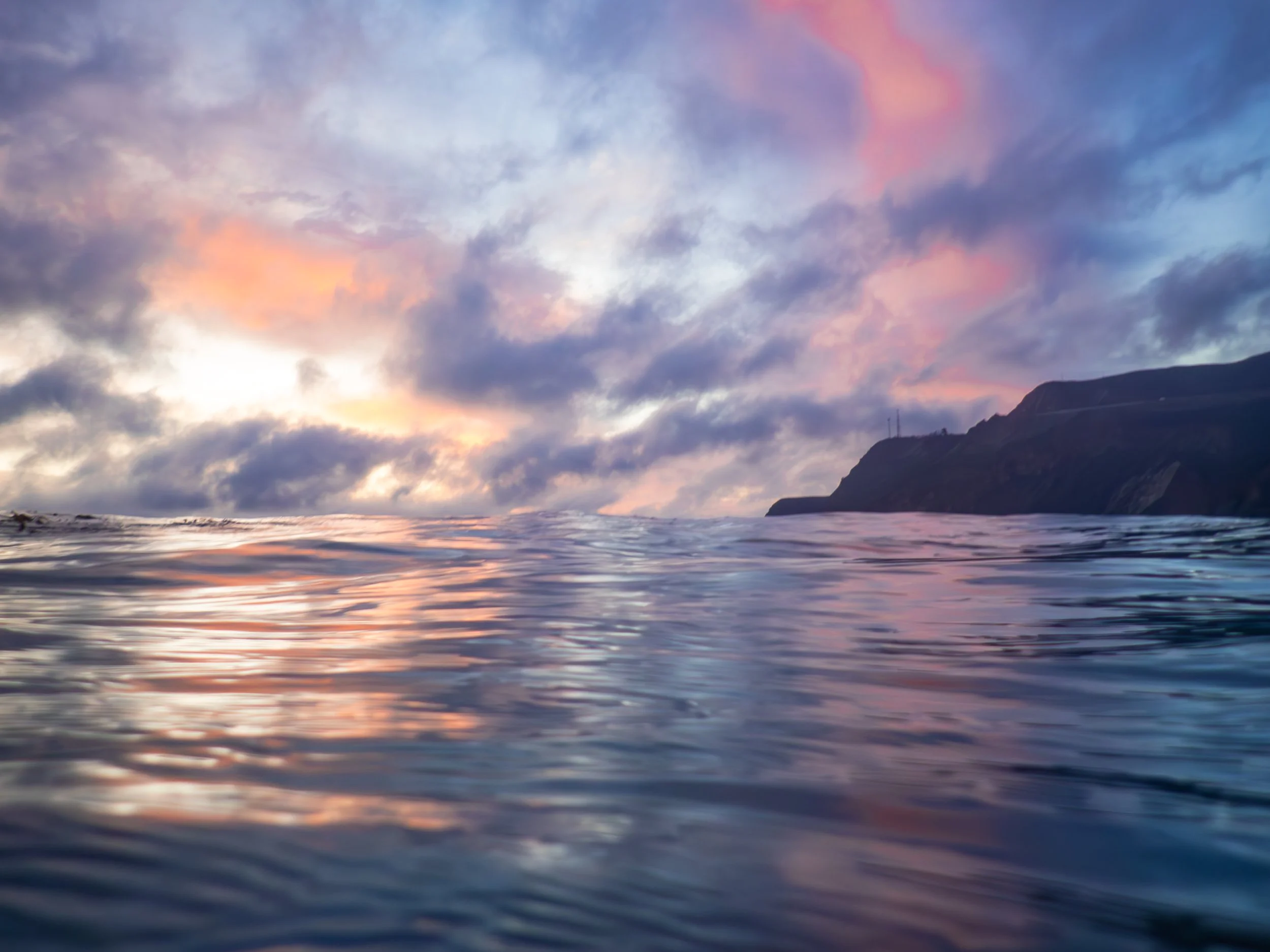 Ocean water reflecting a colorful sky with pink and purple clouds near a silhouetted mountainous coastline at sunset.