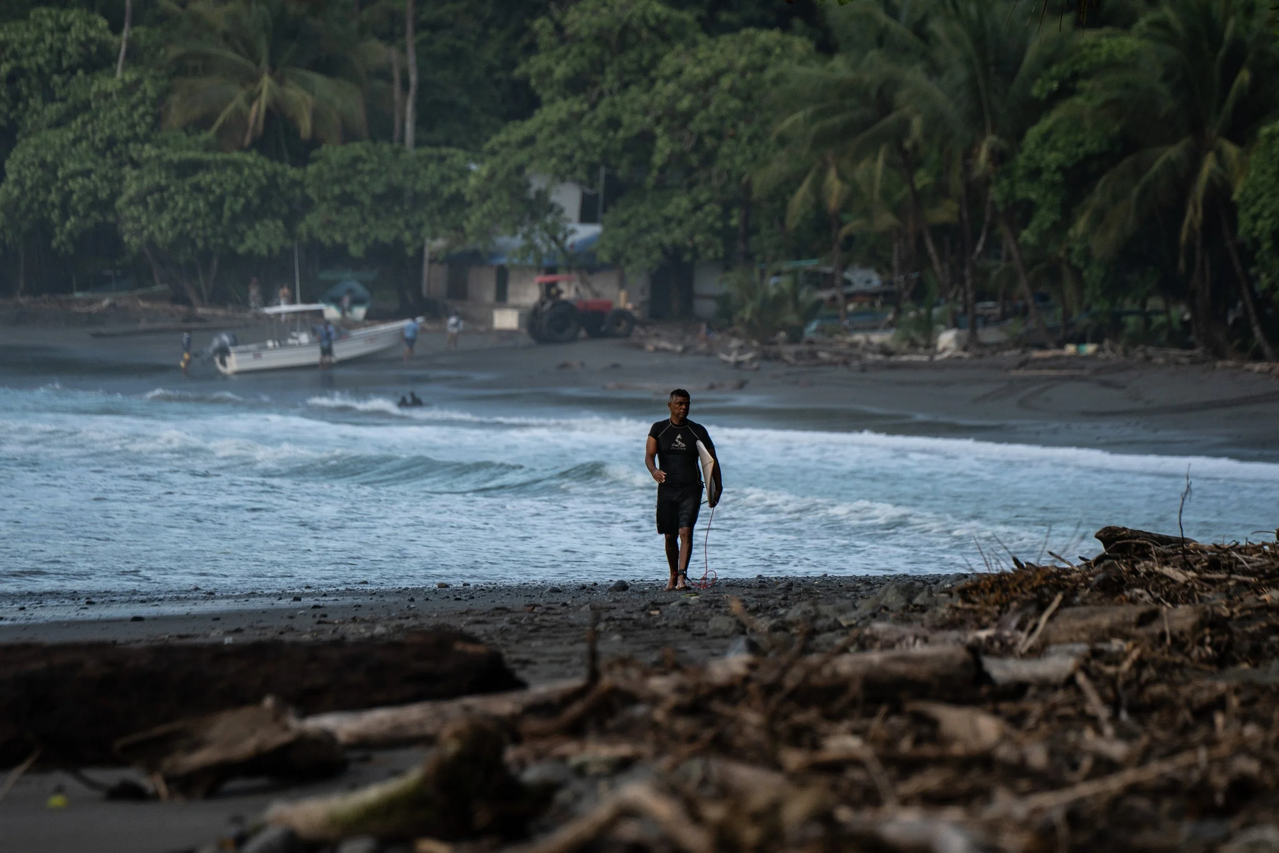 A man walking along the shoreline near the water with a surfboard under his arm, surrounded by trees and houses, during dusk or dawn.