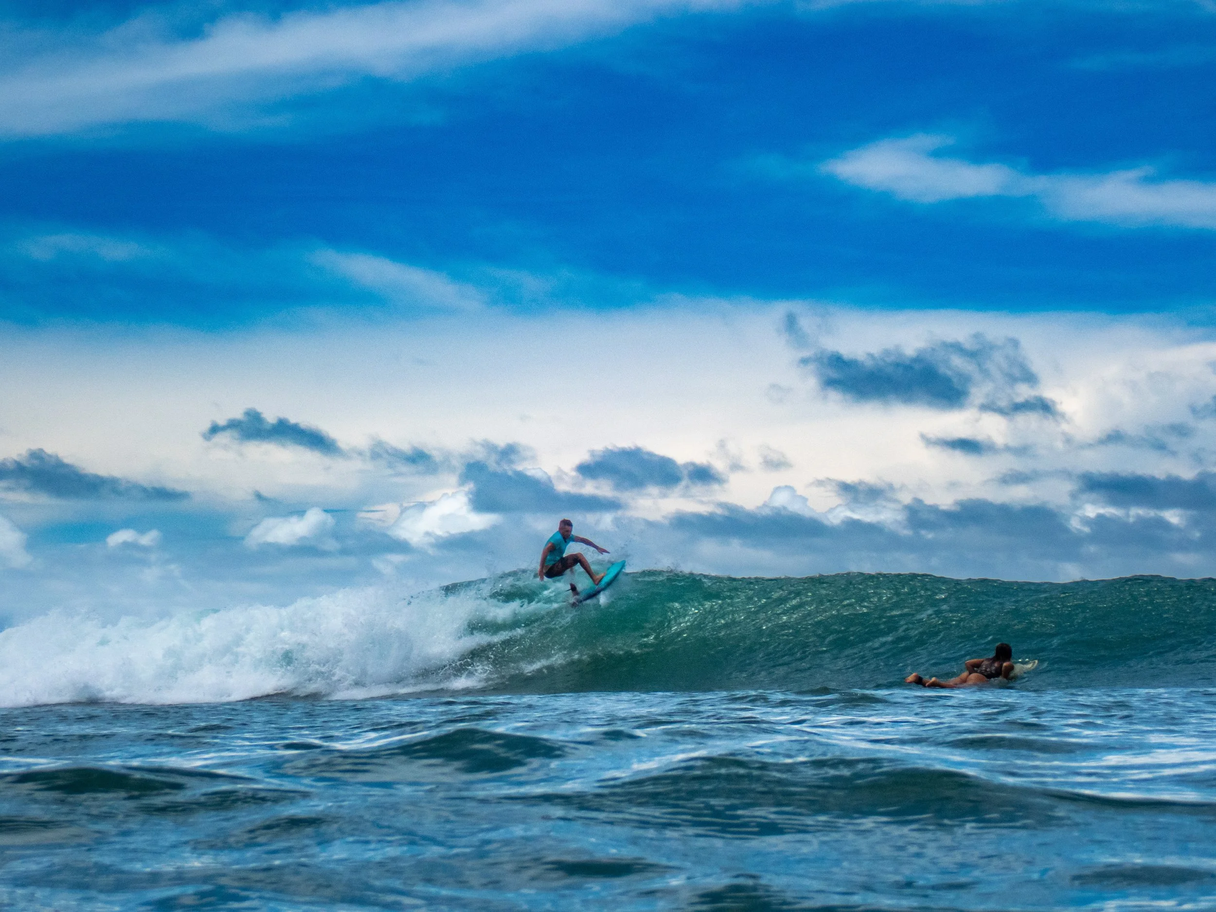 A person surfing on a wave in the ocean, with another person lying on a surfboard in the water nearby under a partly cloudy sky.