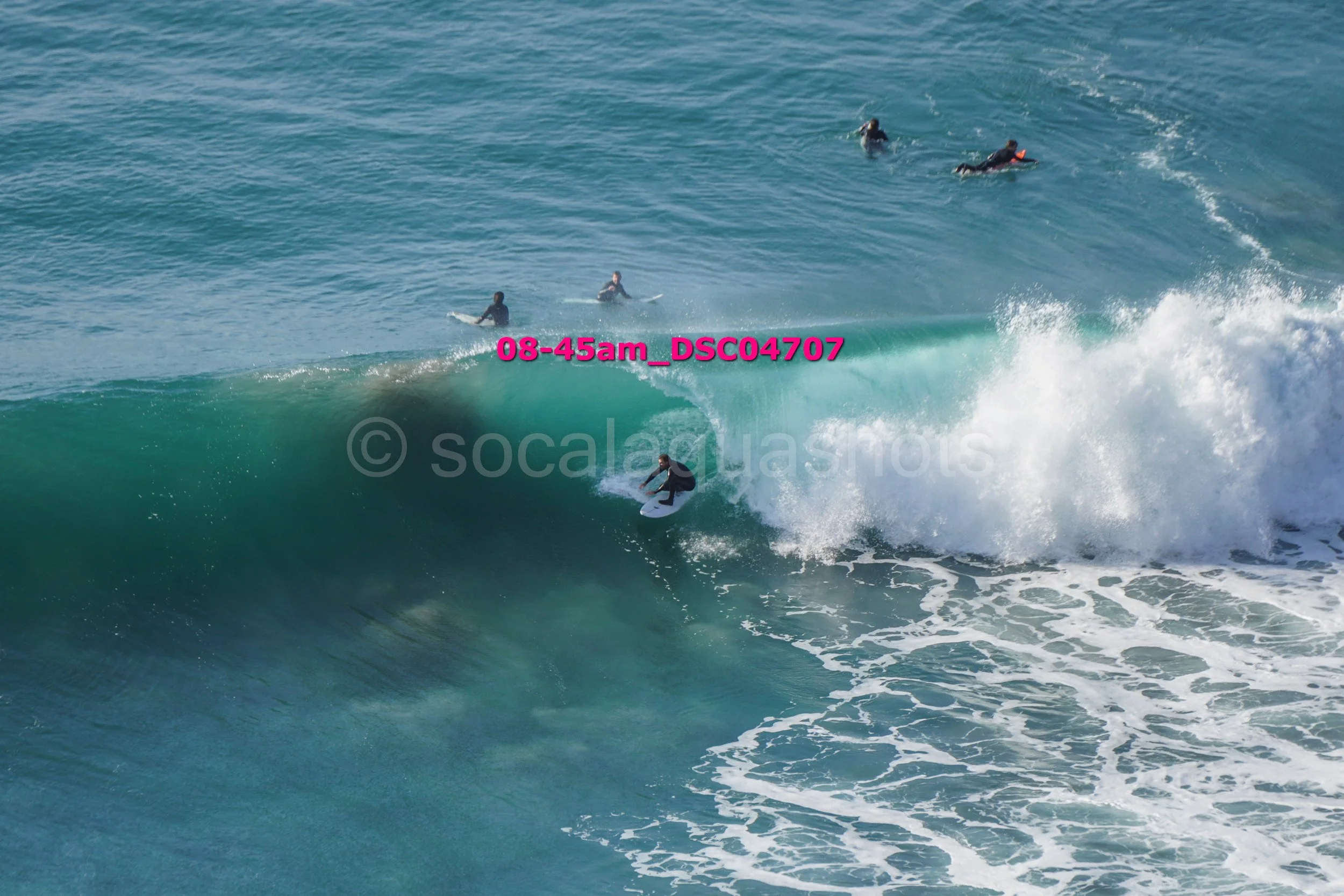 Surfer riding a large wave while several people swim and surf in the background in the ocean.