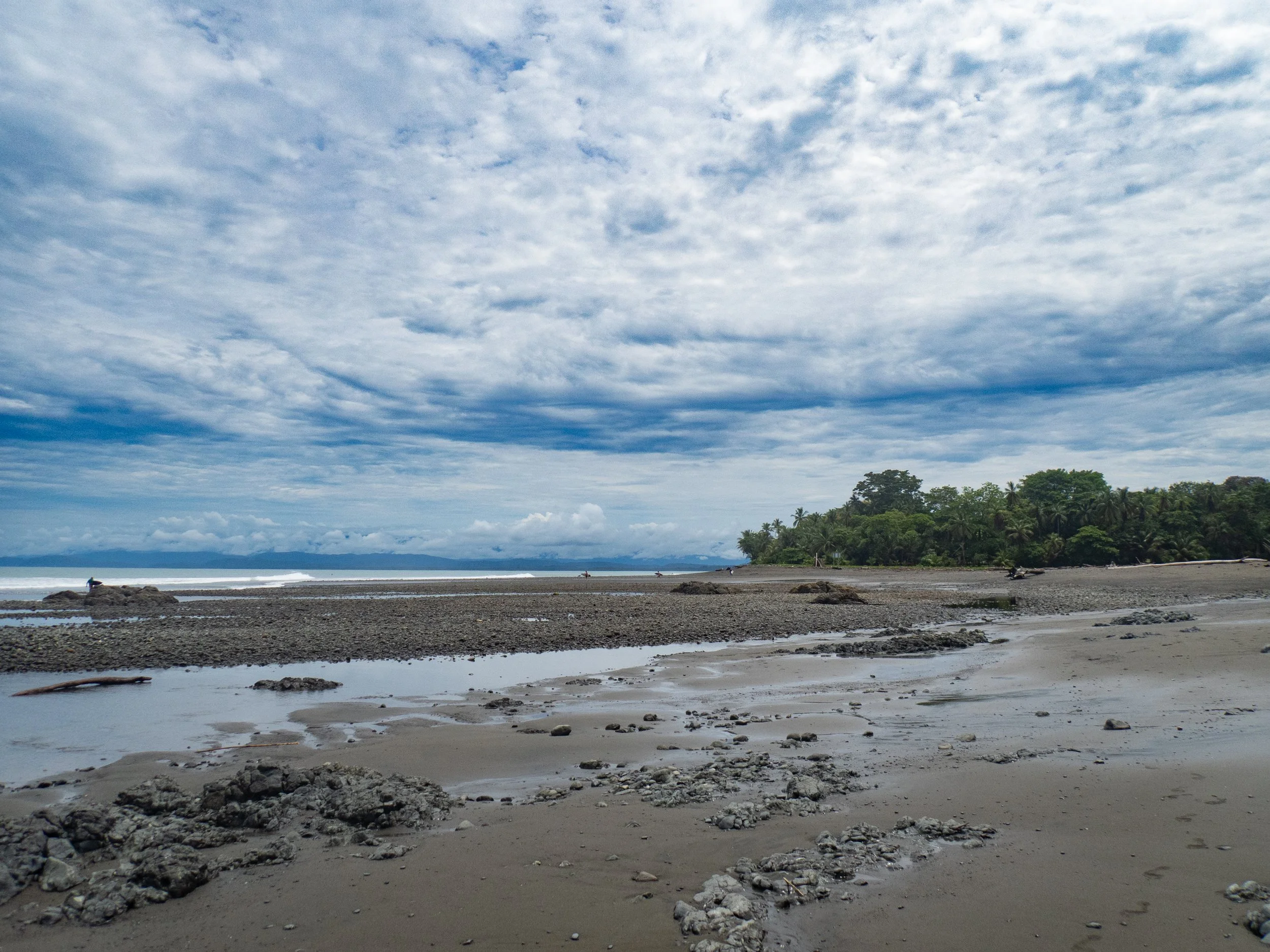 A sandy beach with scattered rocks and shallow pools of water, backed by green trees and a partly cloudy sky.