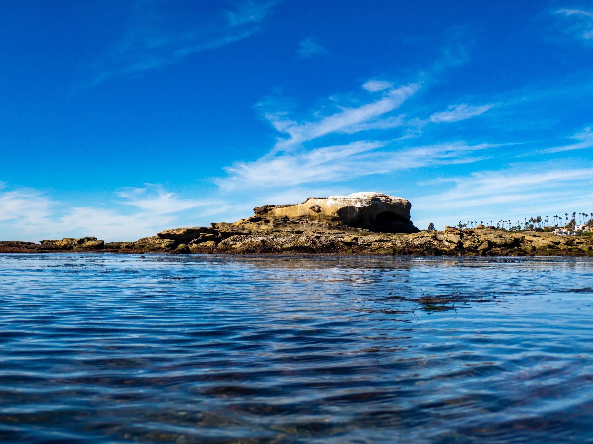A coastal scene with a large rock formation in the water, clear blue sky with wispy clouds, and some palm trees in the distance.