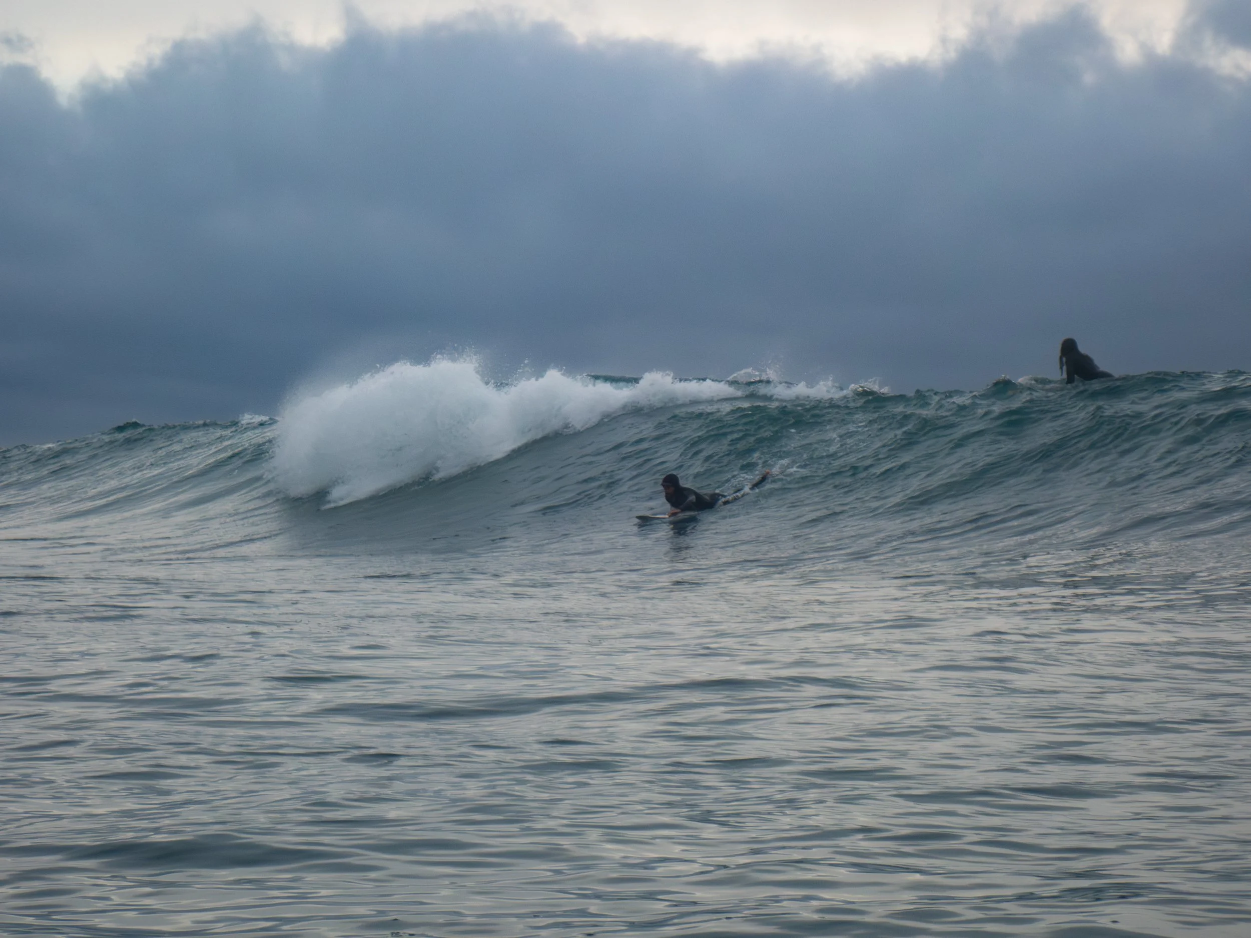 Surfers in wetsuits riding and waiting on ocean waves under a cloudy sky.