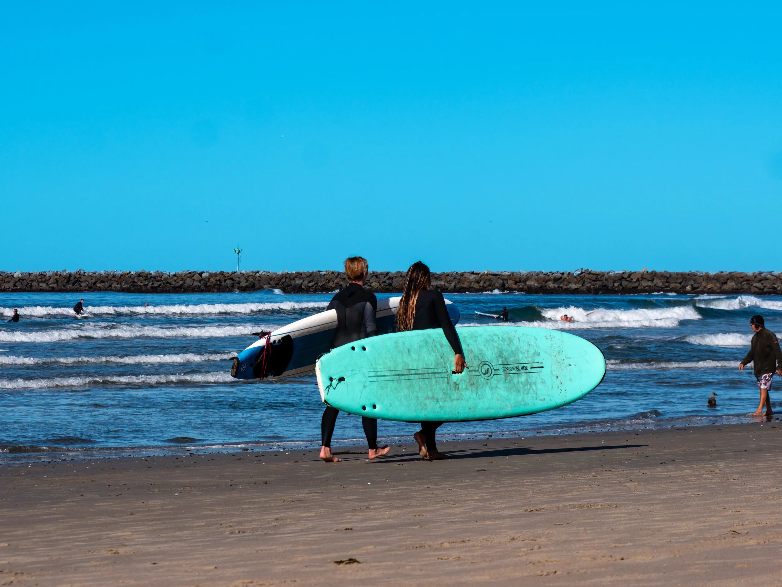 Two surfers walking on the beach holding surfboards, with people surfing in the ocean and a rocky breakwater in the background.