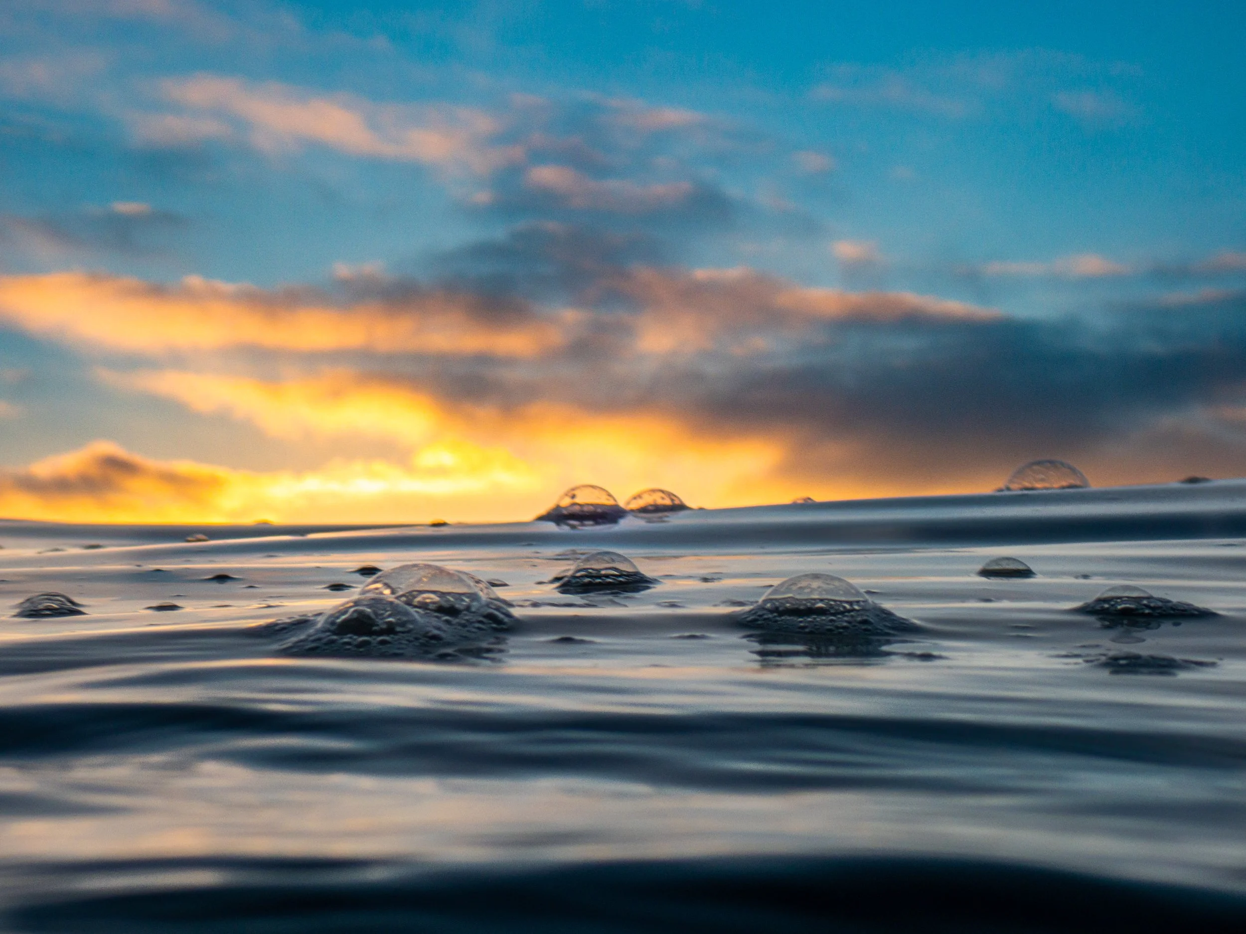 Close-up view of water with bubbles on the surface, under a colorful sky with clouds during sunset or sunrise.