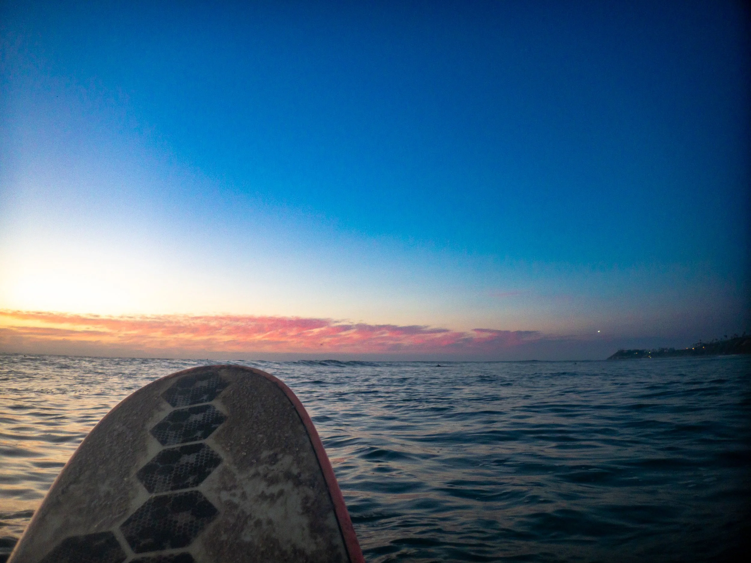 View of the ocean during sunset with a surfboard in the foreground and a coastline in the distance.