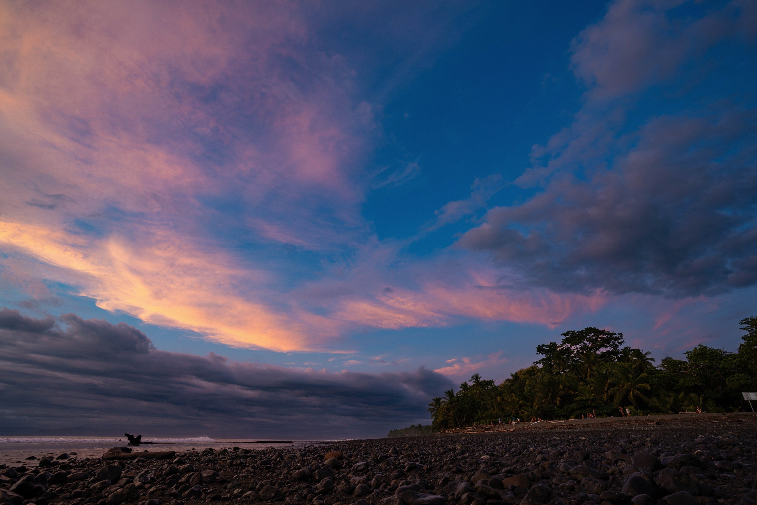 A colorful sunset sky over a beach with dark rocks and silhouetted trees, with some people in the distance on the shoreline.