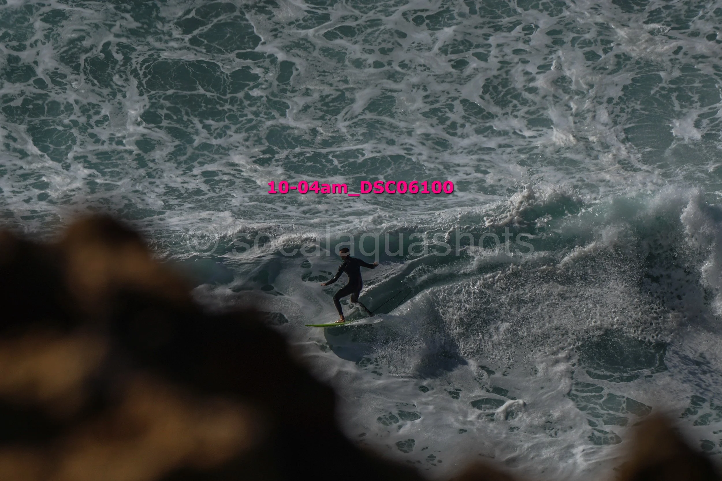 A person surfing on a wave at the ocean, viewed from behind through some dark rocks or plants in the foreground.
