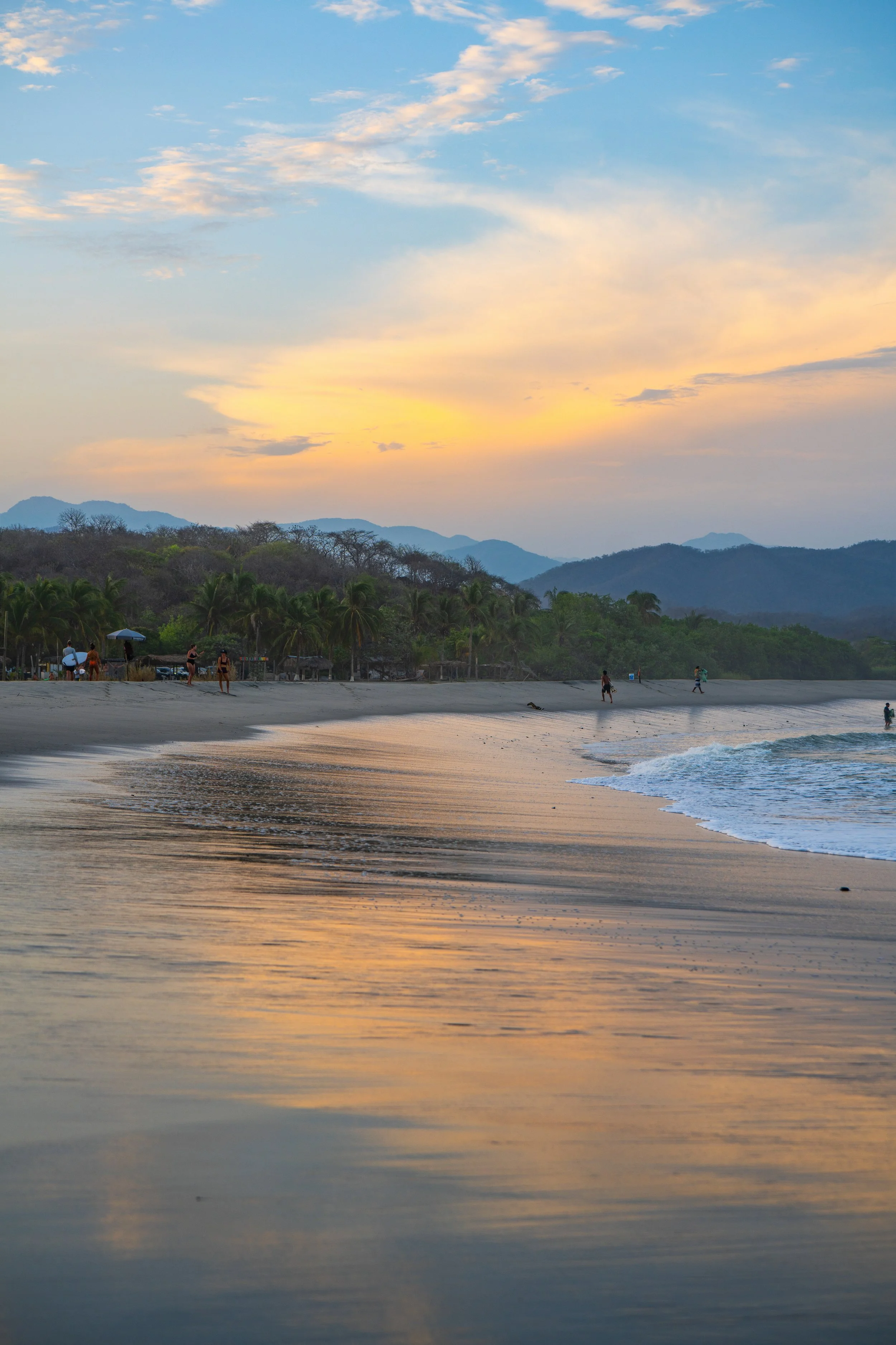 Sunset over a beach with mountains in the background, some people walking along the shoreline, palm trees, and a partly cloudy sky.