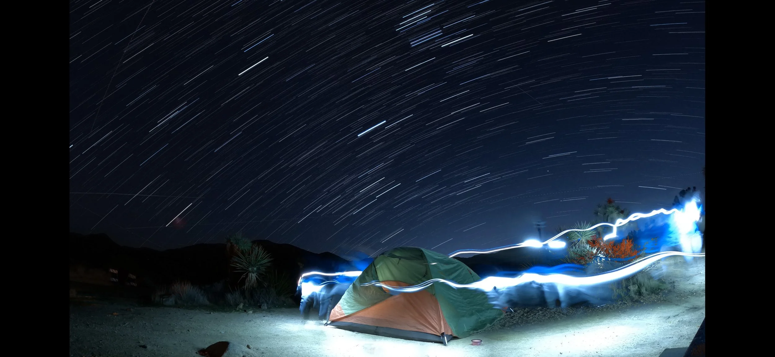 Long exposure photograph of a starry night sky over a desert campsite with a tent, light trails from people moving around, and star trails showing circular motion.