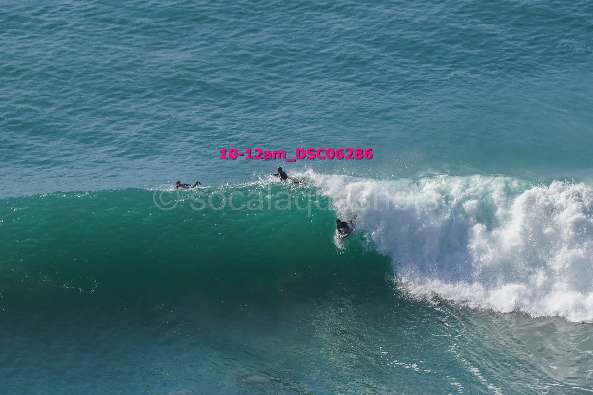 Three surfers riding a large ocean wave with one falling off, captured during daylight.