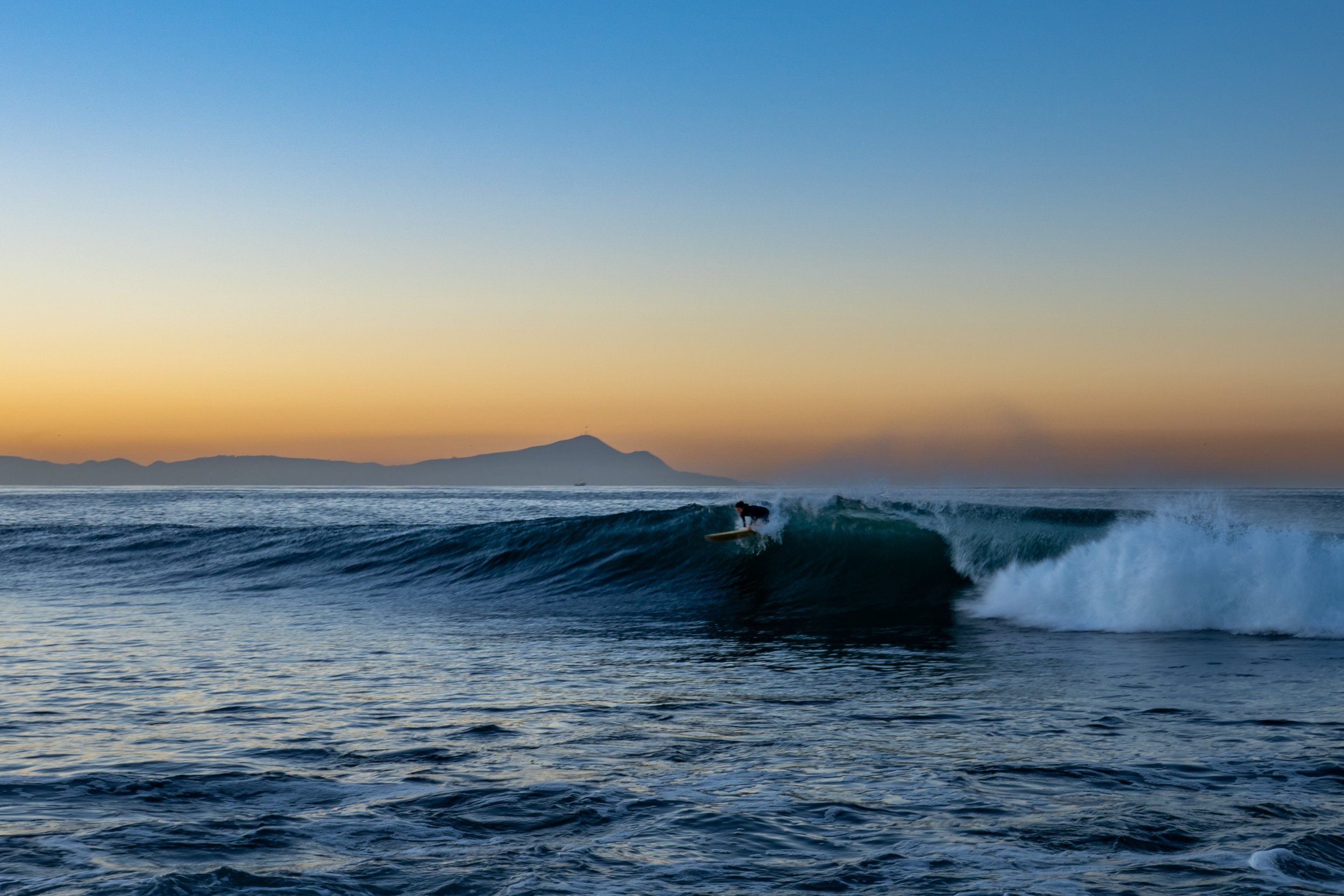 A person surfing on a wave during sunset with a mountainous landscape in the background.