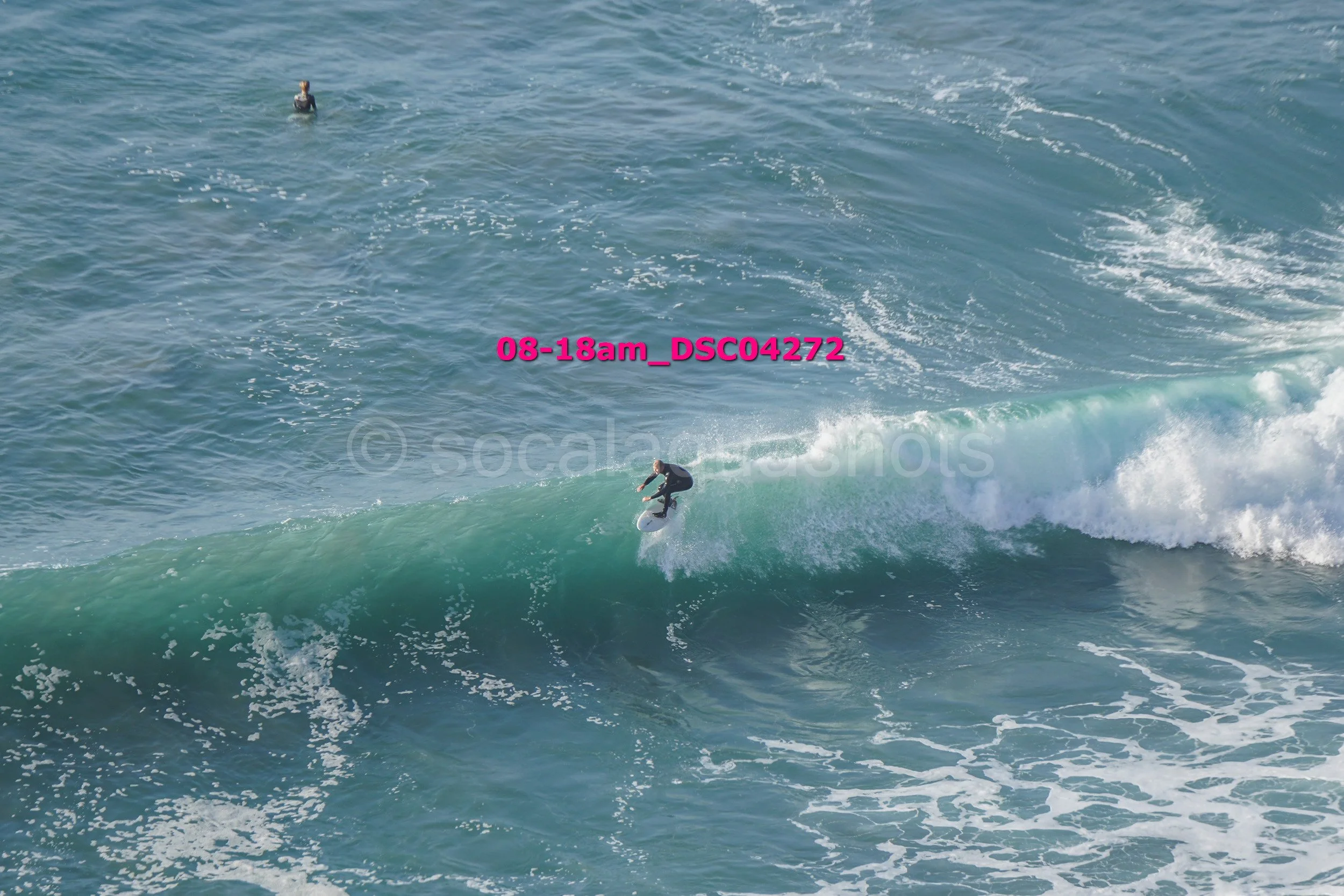Surfer riding a wave in the ocean, with a person swimming in the background.