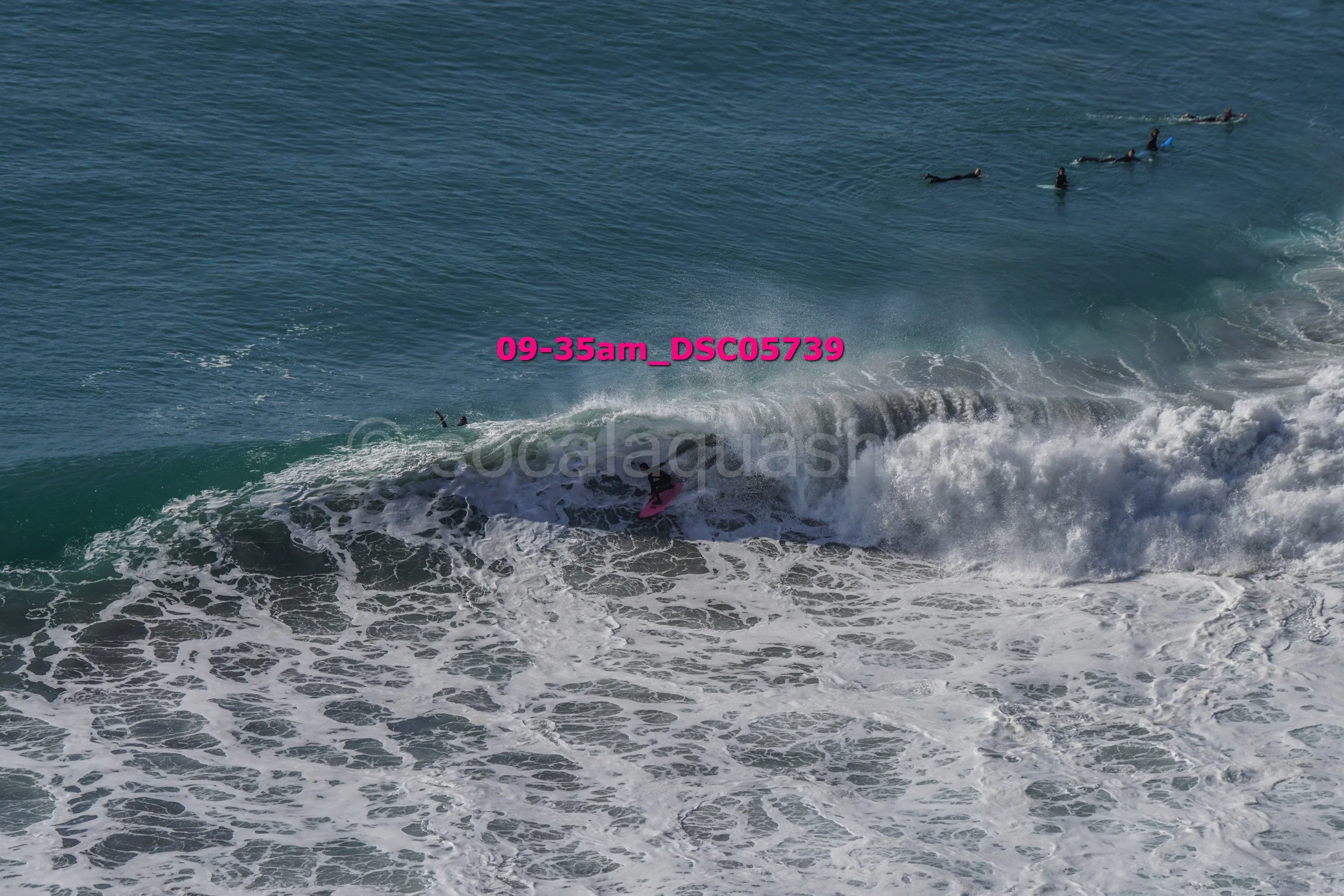 Surfer riding a wave in the ocean with swimmers visible in the background.